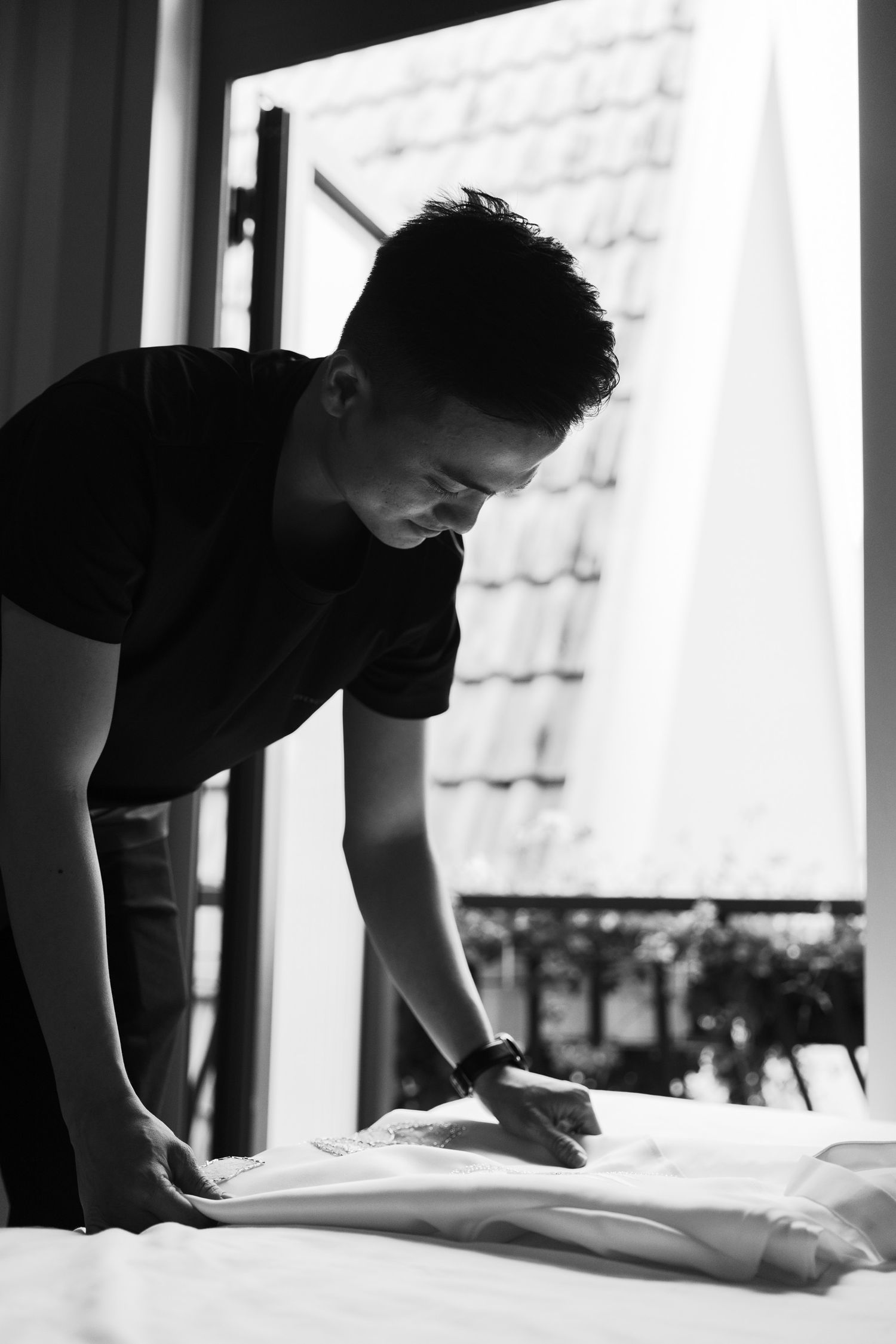 Black and white photo of a person making a bed in a room with natural light.