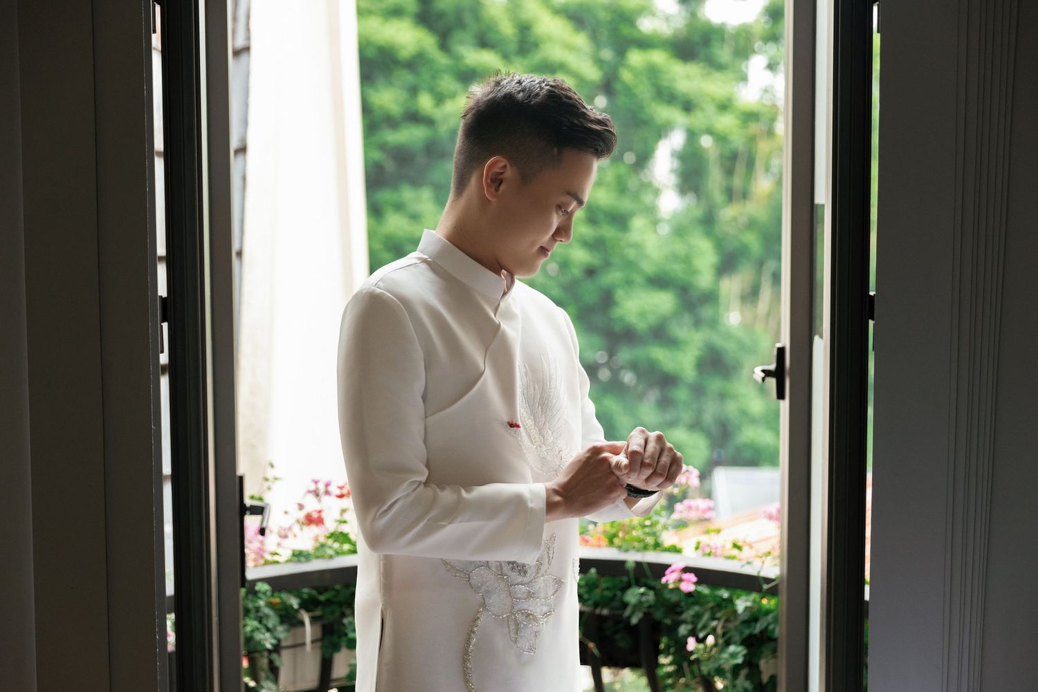 Individual in white traditional attire looking at phone by window with greenery outside.