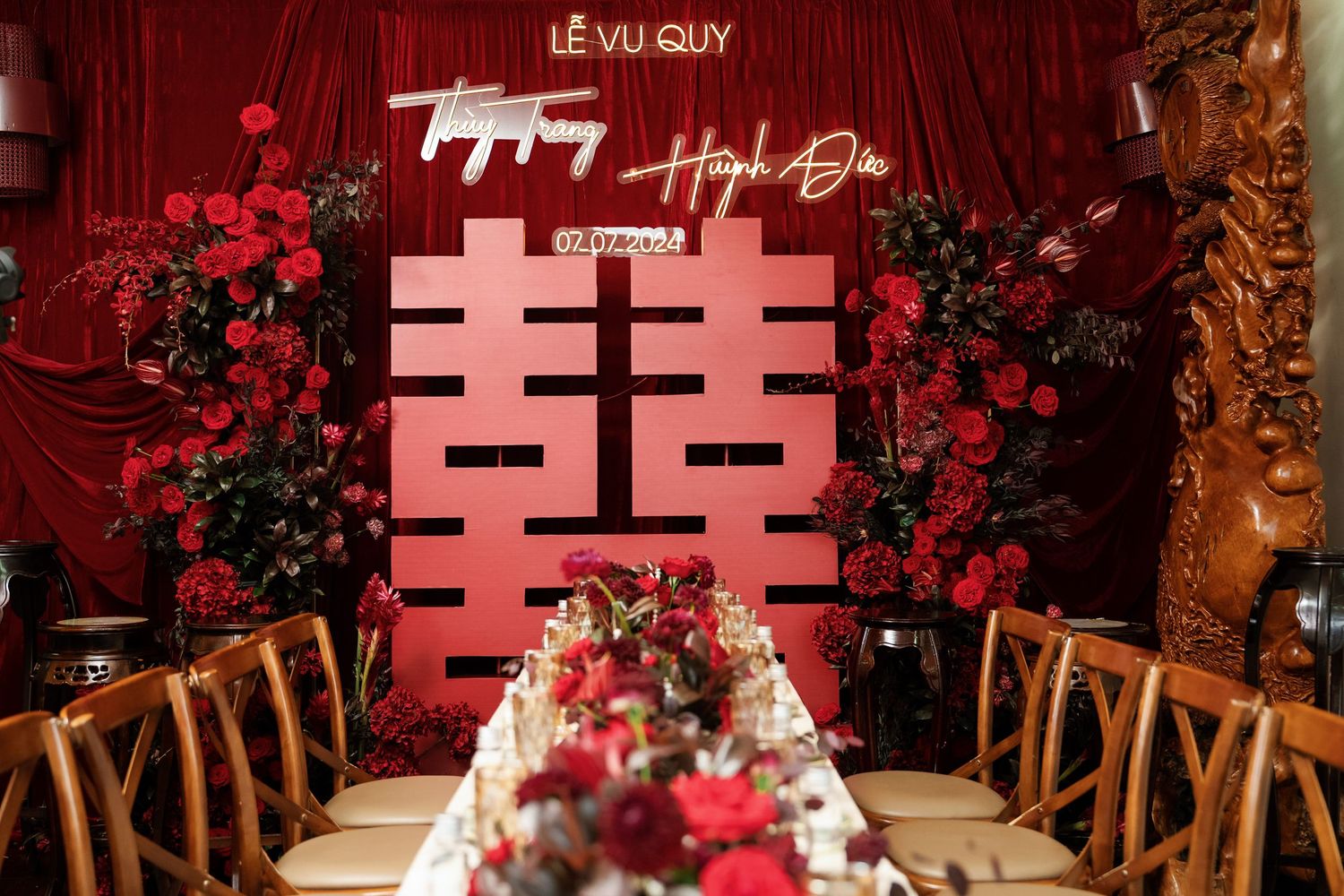 Wedding reception setup with red double happiness symbol and wooden chairs around long table.
