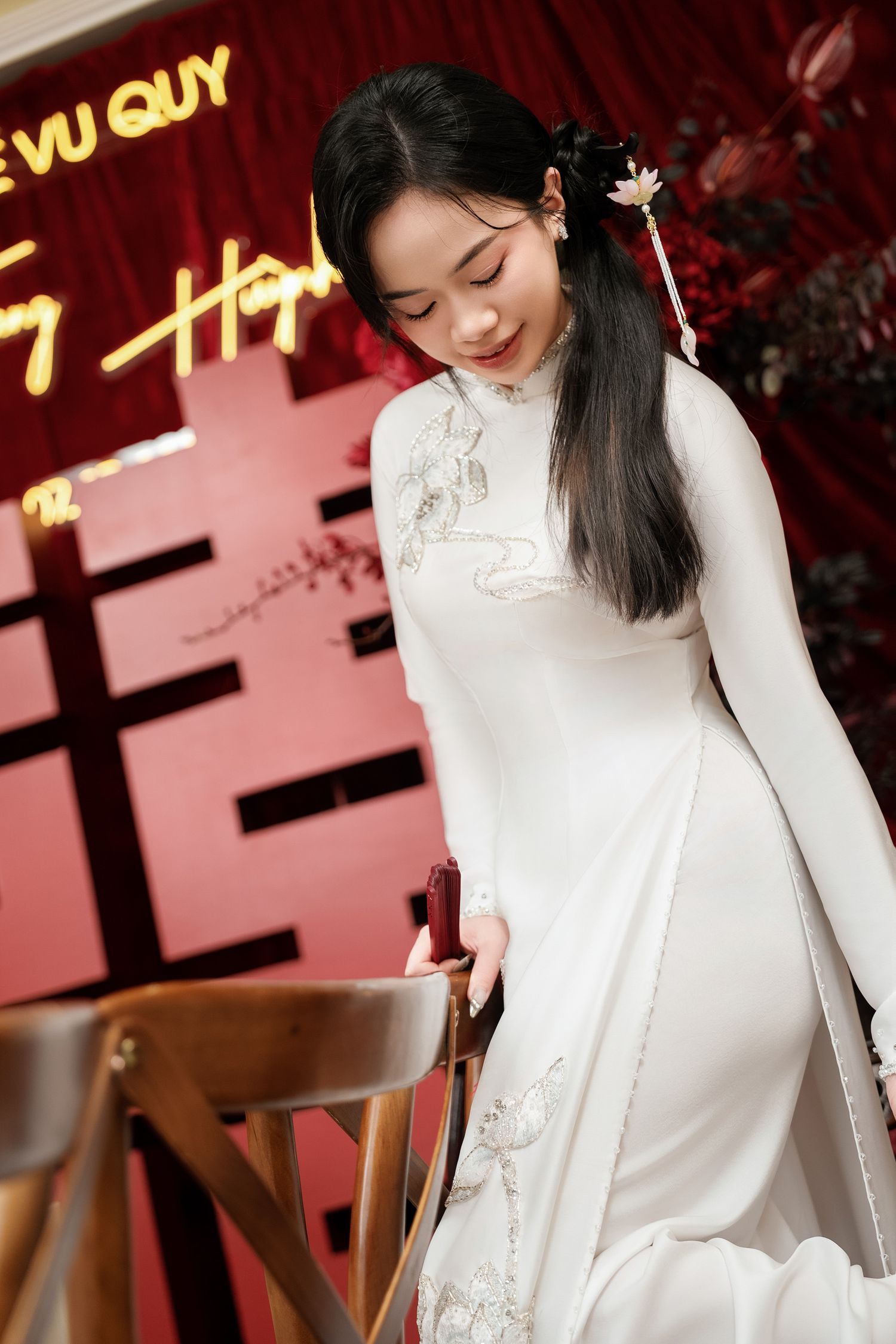 Person in elegant white traditional ao dai dress poses against red backdrop with decorative signage.