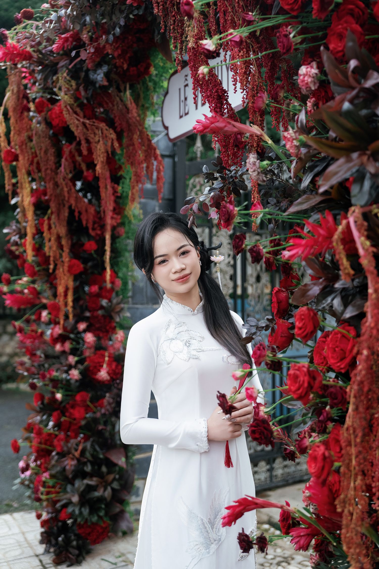 Person in white ao dai poses among vibrant red floral decorations outdoors.