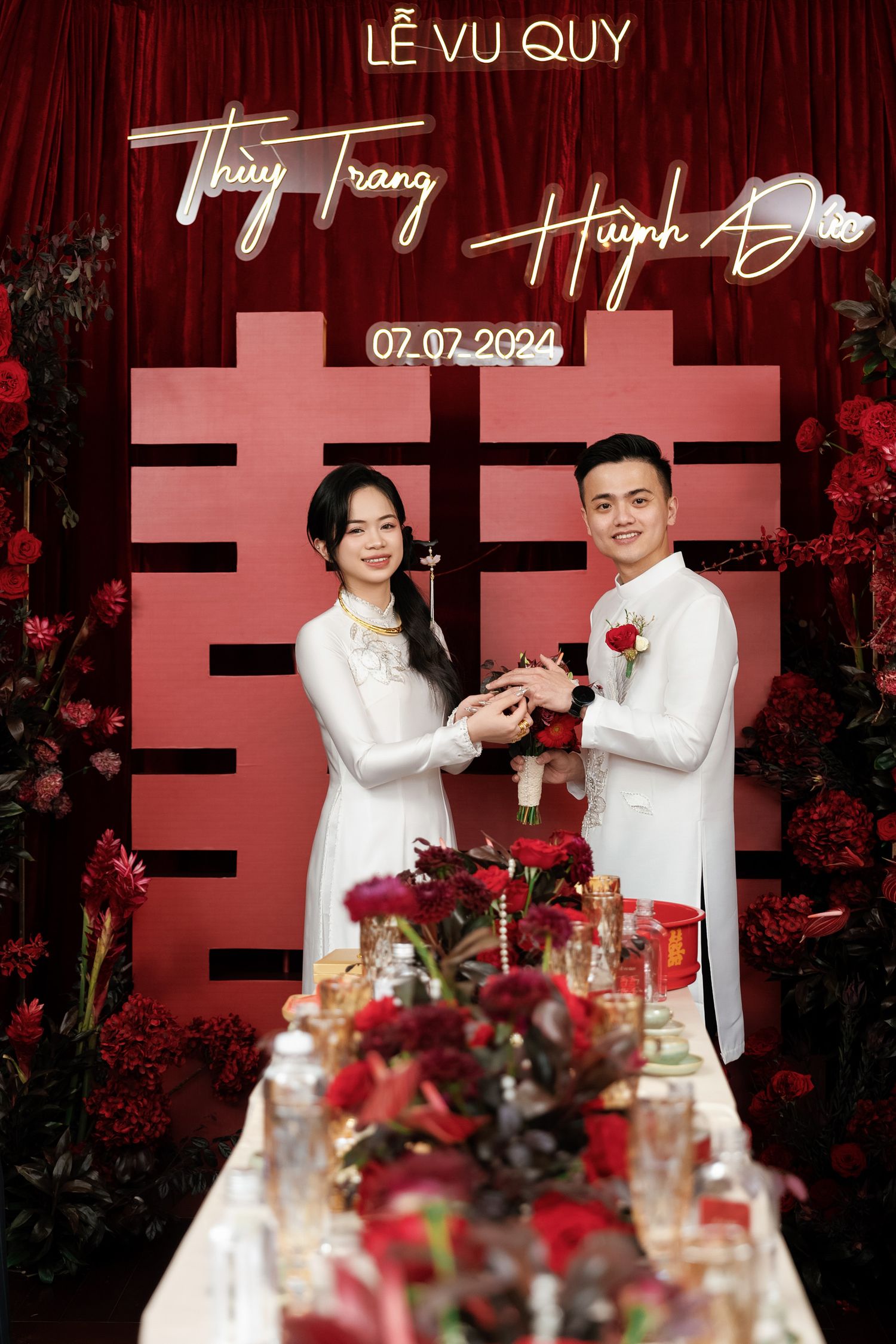 Wedding couple in white ao dai attire pose with red floral decorations and double happiness symbol.