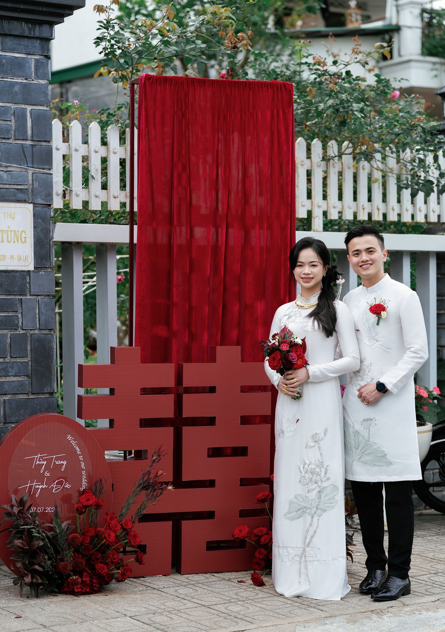Couple in traditional white ao dai pose by red double happiness symbol and white picket fence.
