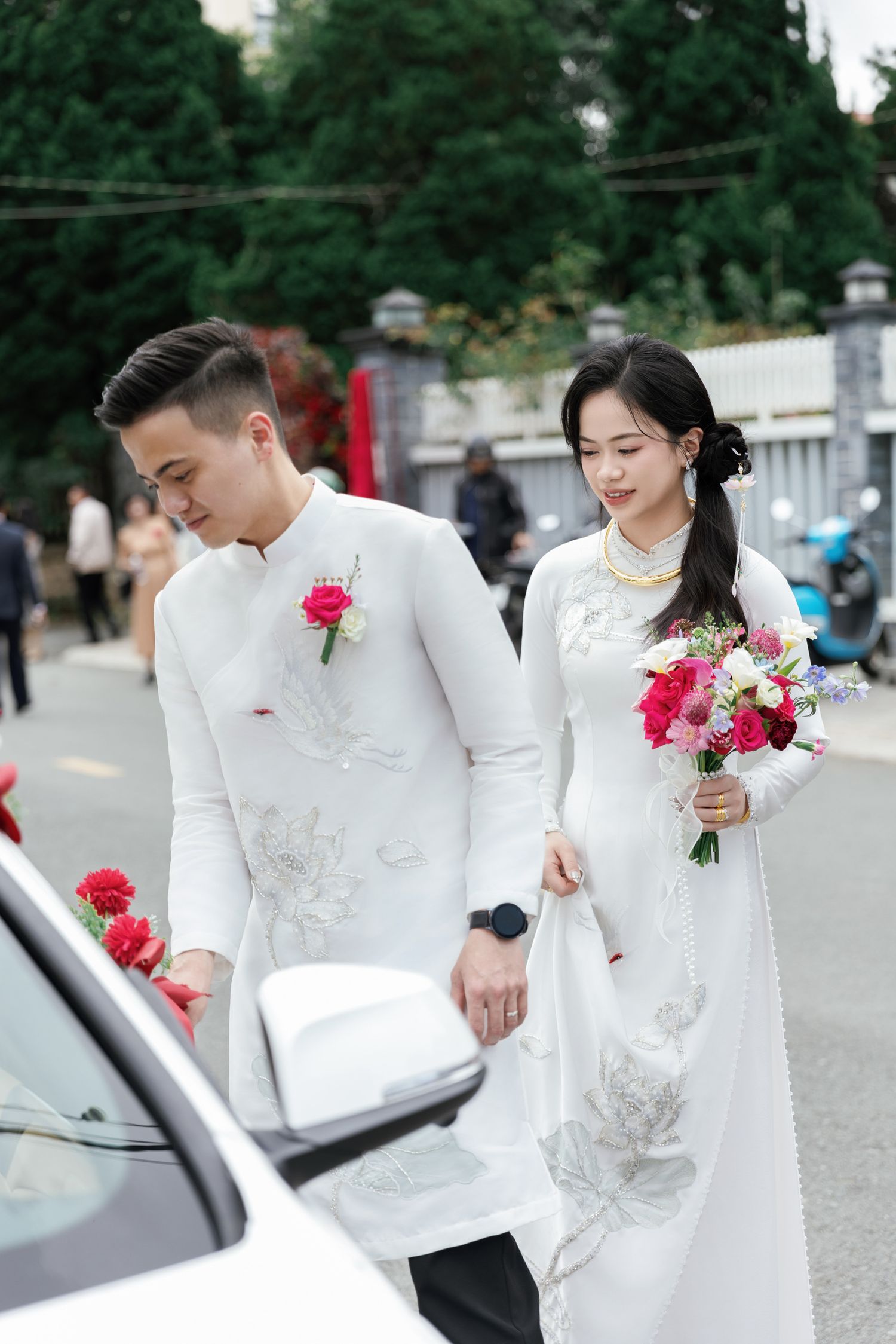 A couple in traditional white ao dai attire walking beside their wedding car.