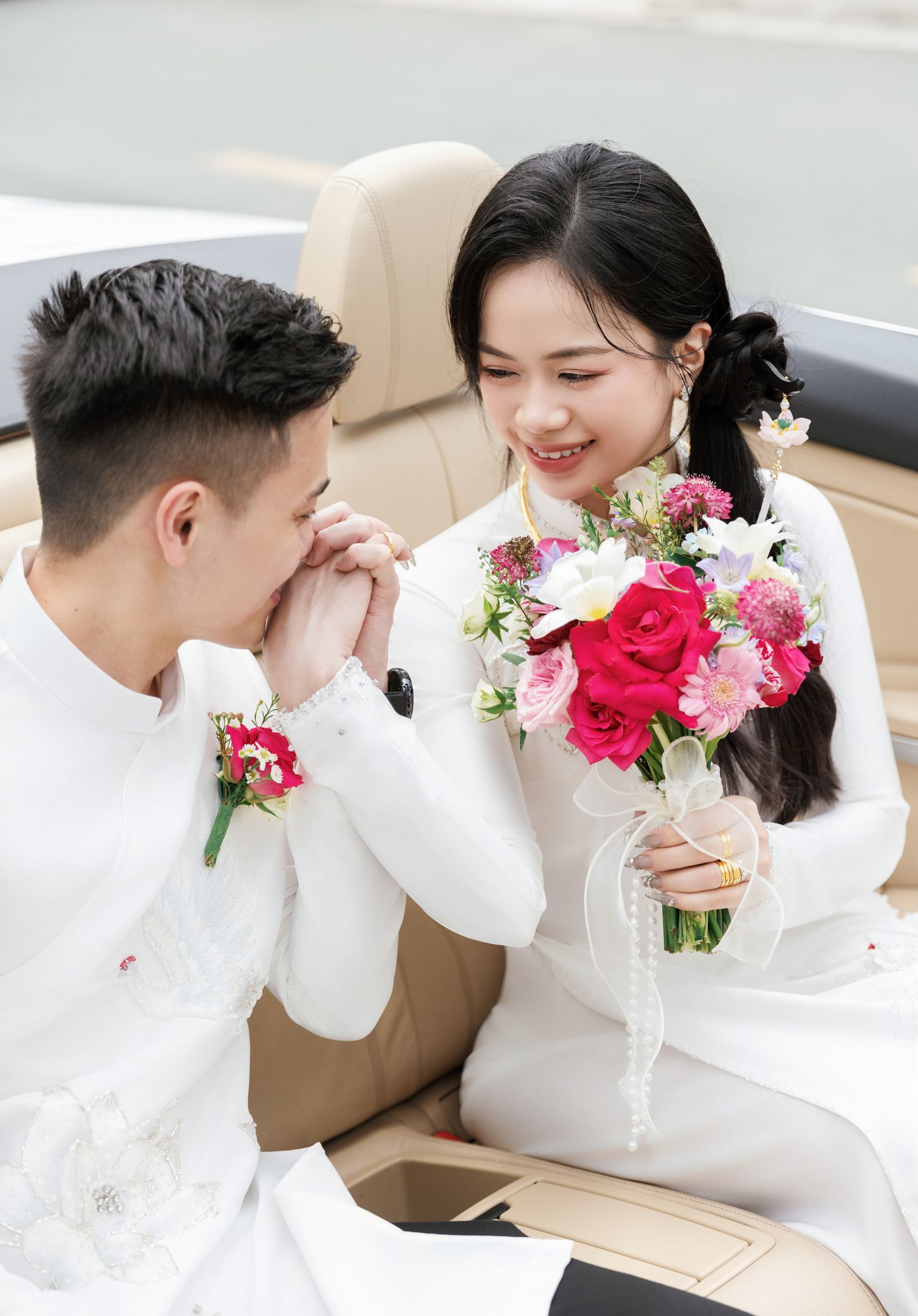 Tender moment between couple in white traditional attire inside car with pink bouquet.