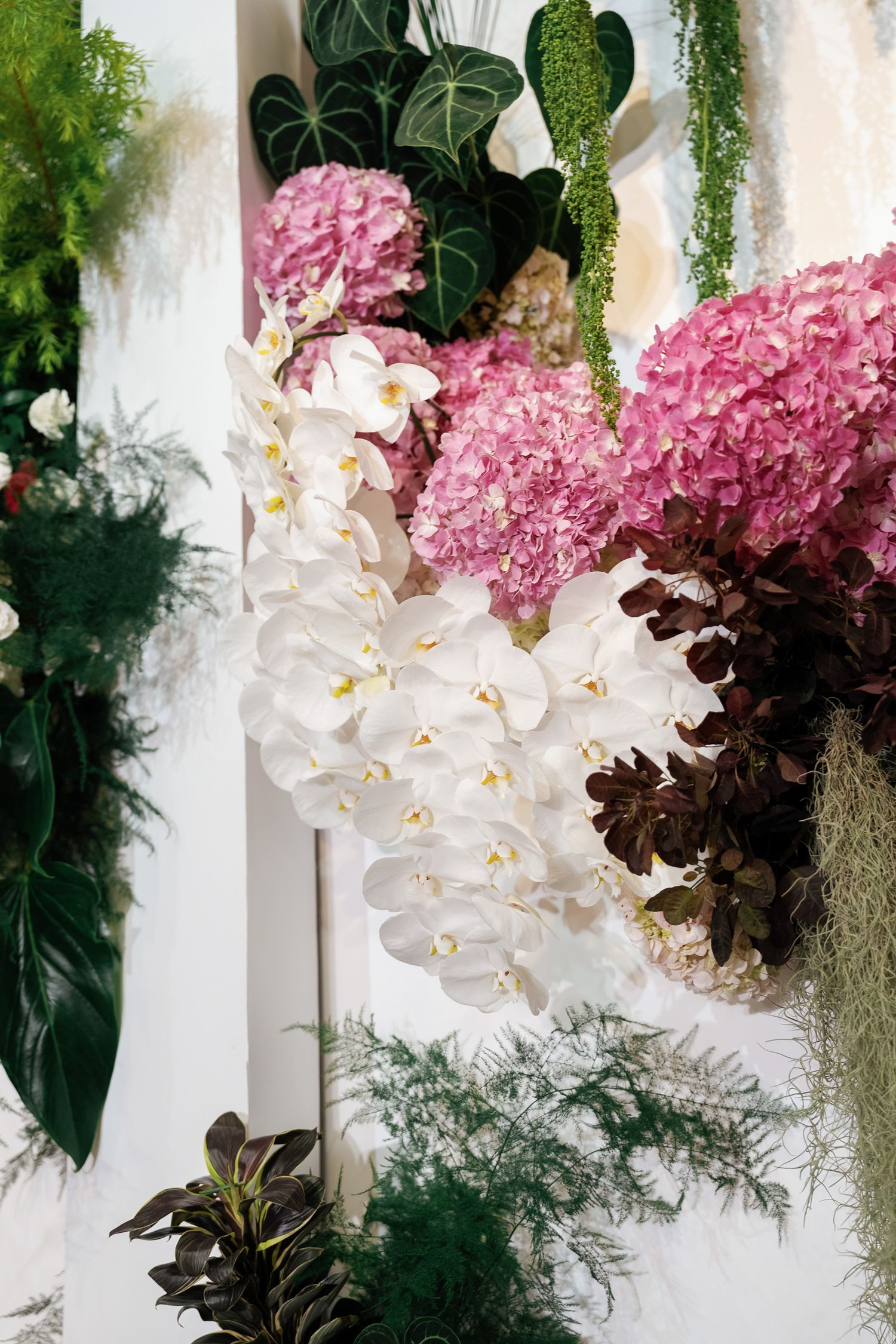 White orchids and pink hydrangeas arranged with tropical foliage against white wall.