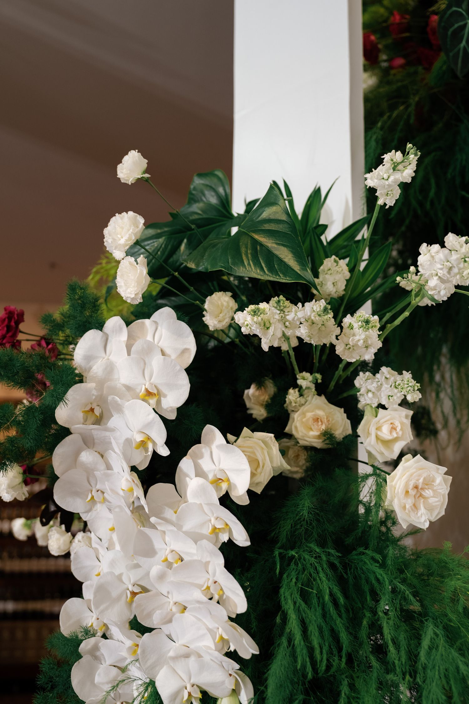 Elegant white orchids and roses arranged with lush green foliage against a white pillar.