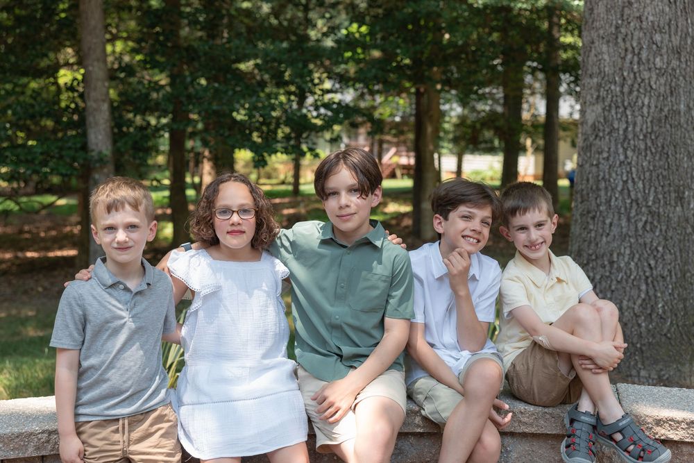 Five children sitting together outdoors on a stone ledge, smiling, surrounded by trees in a park setting.