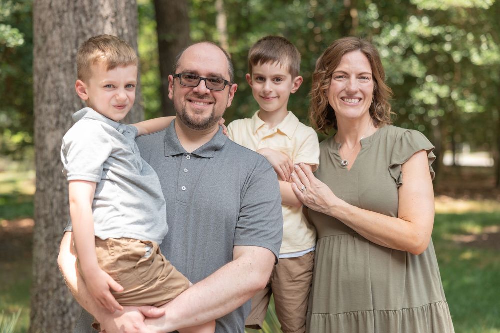 A smiling family of four poses outdoors among trees, with a man holding a young boy beside a woman and older boy.
