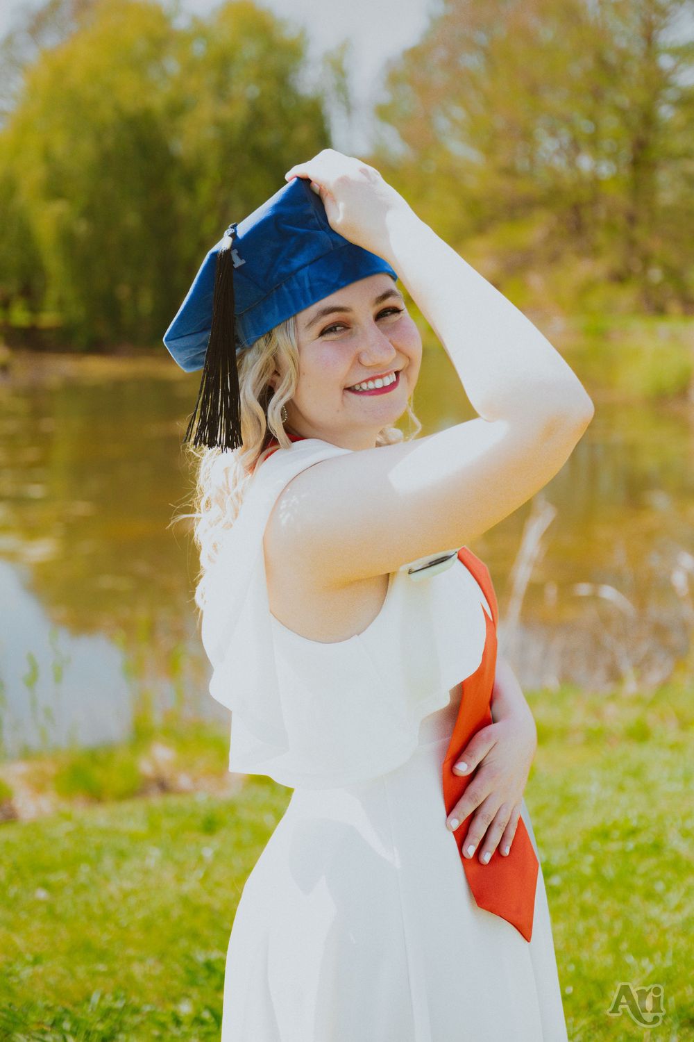 Smiling female graduate in white dress holds blue cap outdoors near pond, wearing orange sash for graduation photos.