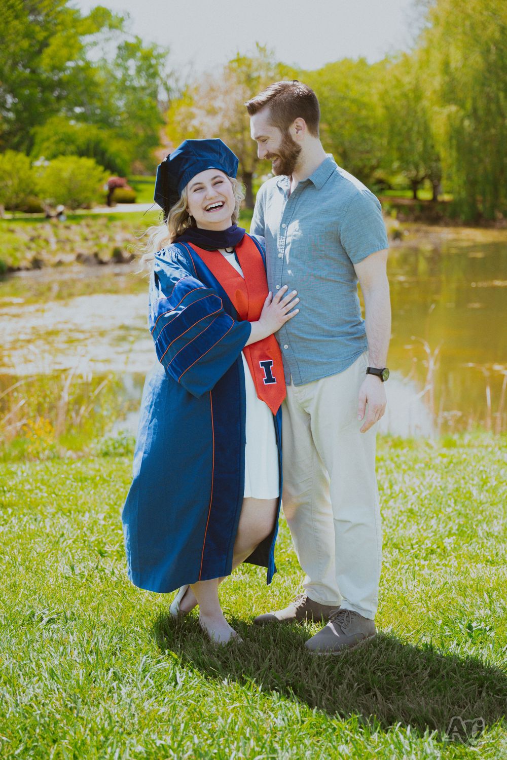 A woman in blue graduation cap and gown laughs with a bearded man outdoors near a pond on a sunny day.