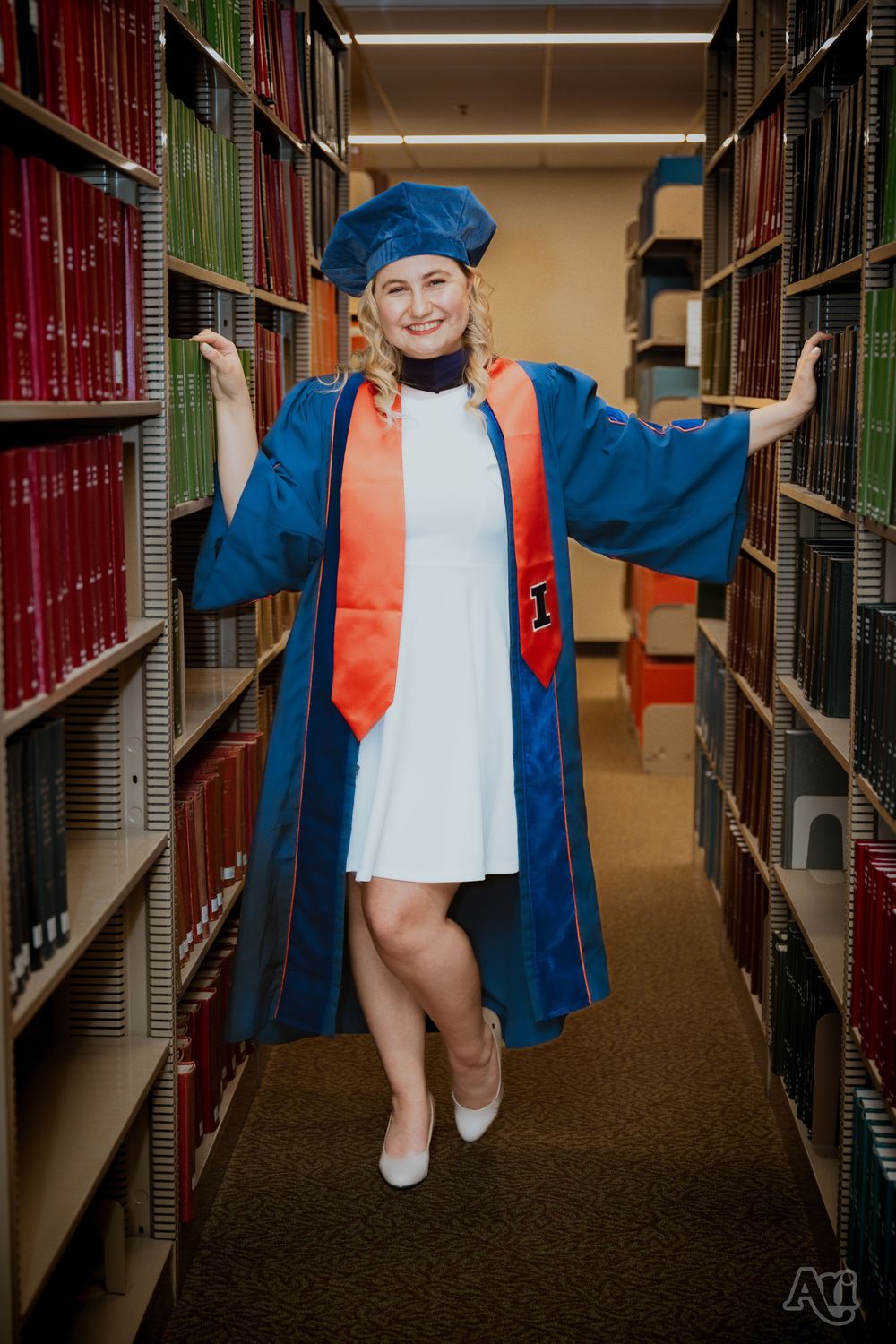 Woman in blue graduation gown and cap poses proudly between library bookshelves wearing an orange stole.