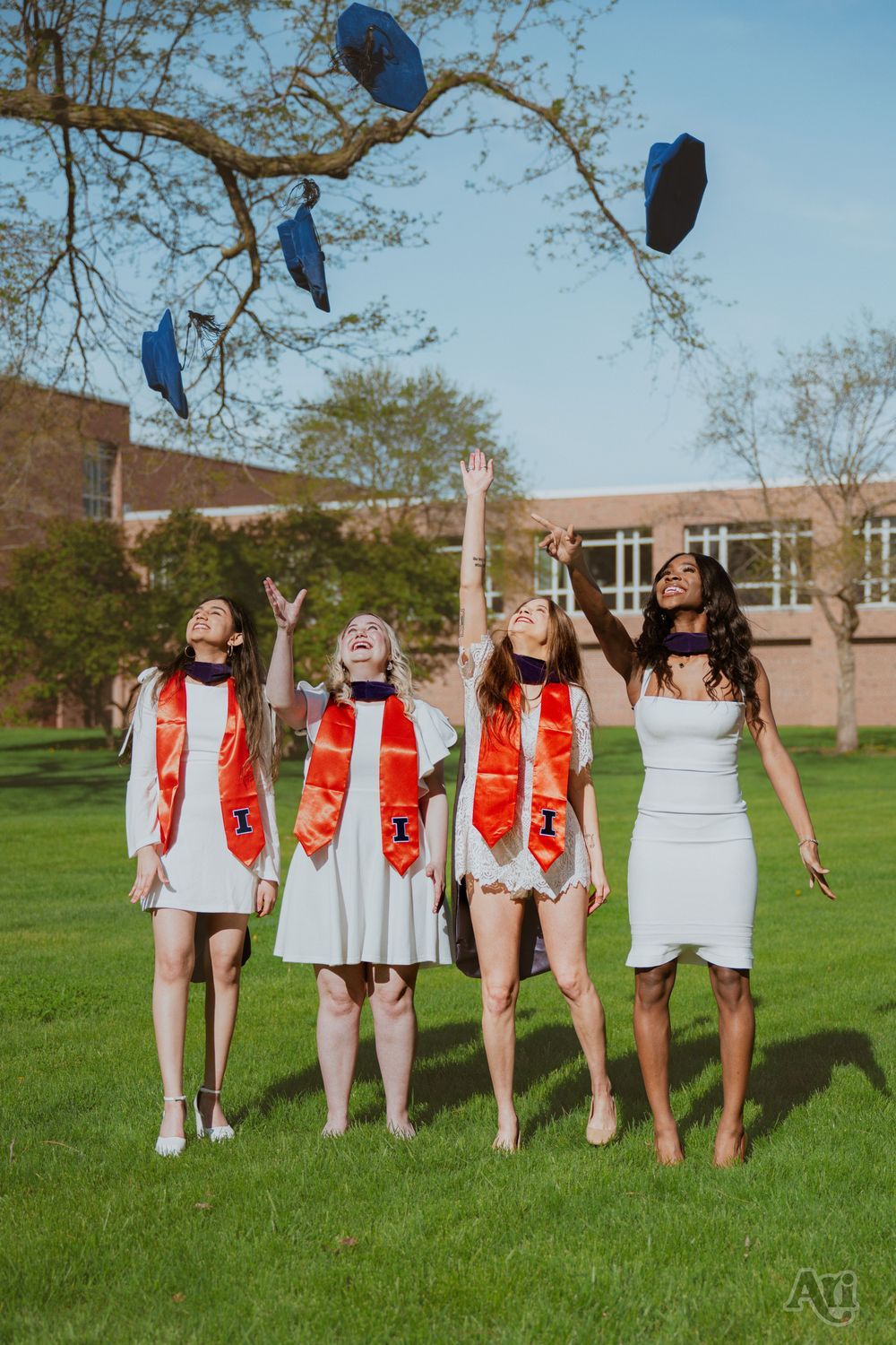 Four college graduates in white dresses and orange stoles toss blue caps in the air outdoors.