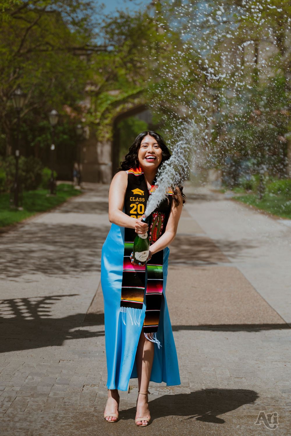 Joyful graduate in blue dress and colorful sash pops champagne bottle outdoors on sunny day.