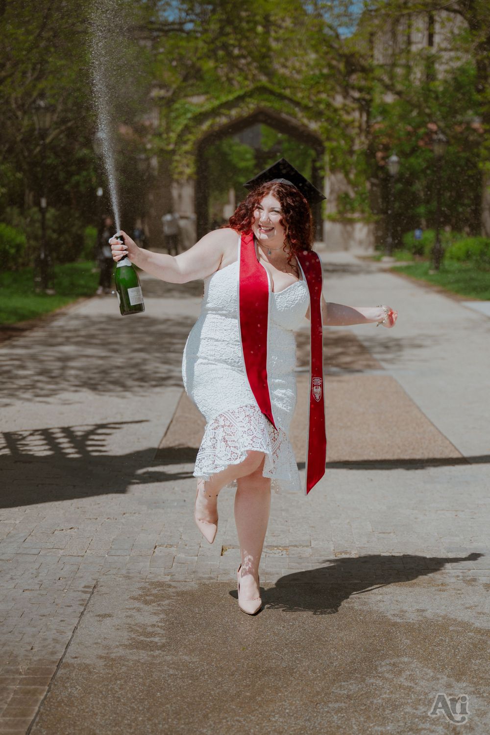 Joyful graduate in white dress and red sash sprays champagne while skipping outdoors on a sunny campus pathway.