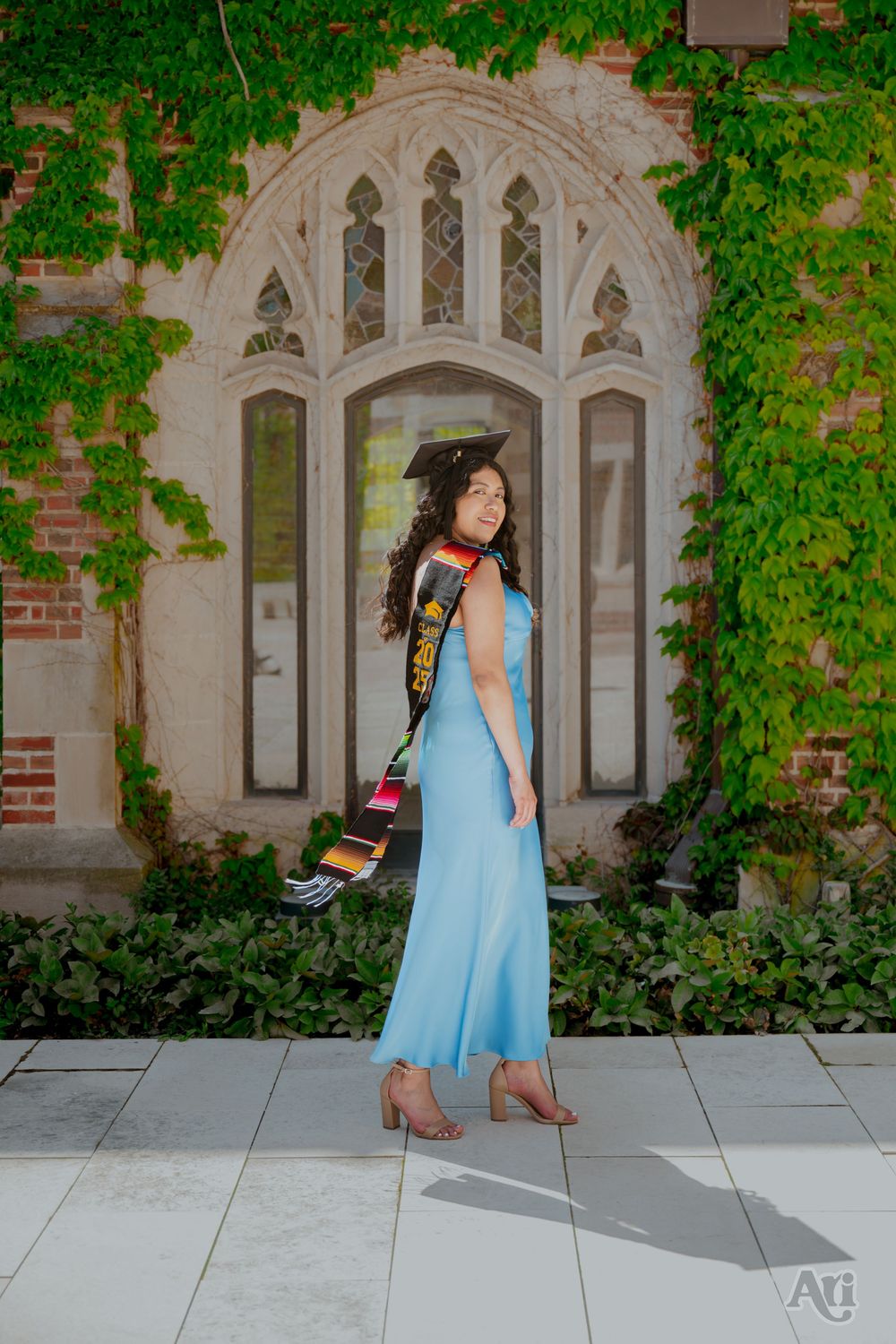A graduate in a light blue gown and cap poses before an ivy-covered stone archway.
