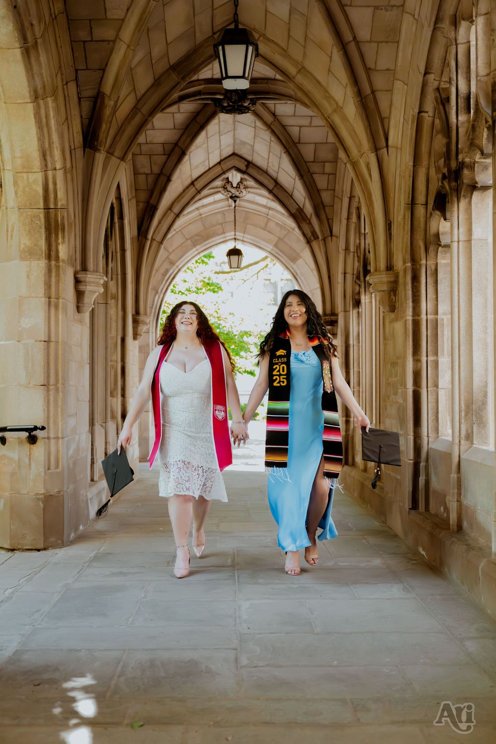 Two graduates in colorful sashes walk hand-in-hand through gothic stone archways celebrating their graduation day.