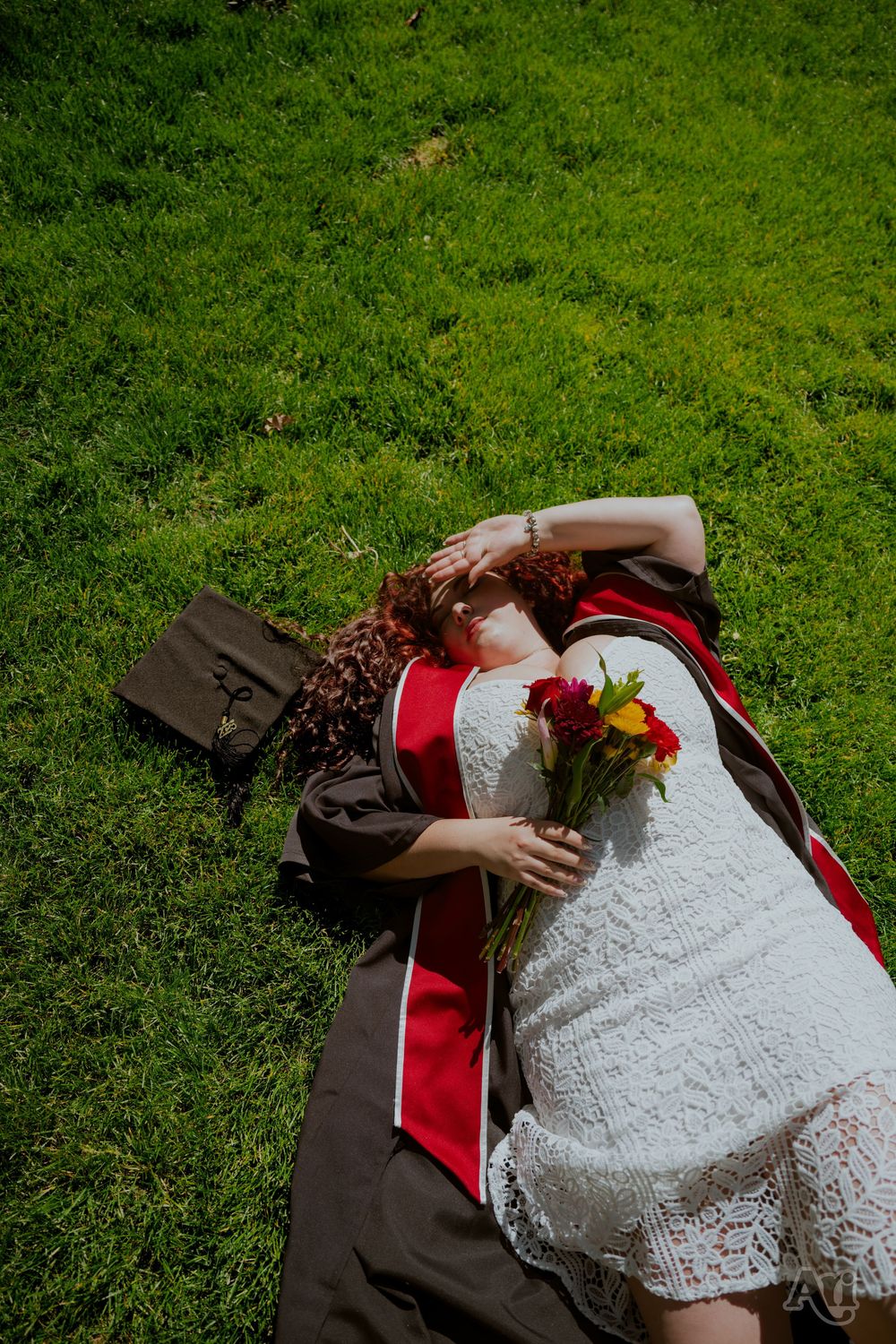 Graduate in red sash lying on green grass holding colorful bouquet with brown bag beside her.