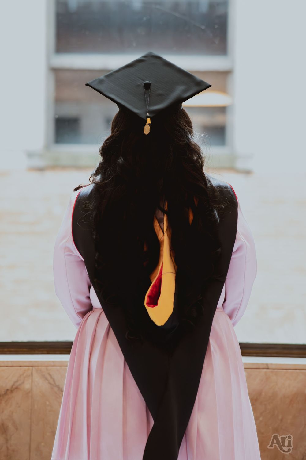 Graduate in pink dress and black cap and gown seen from behind, standing outdoors at graduation ceremony.
