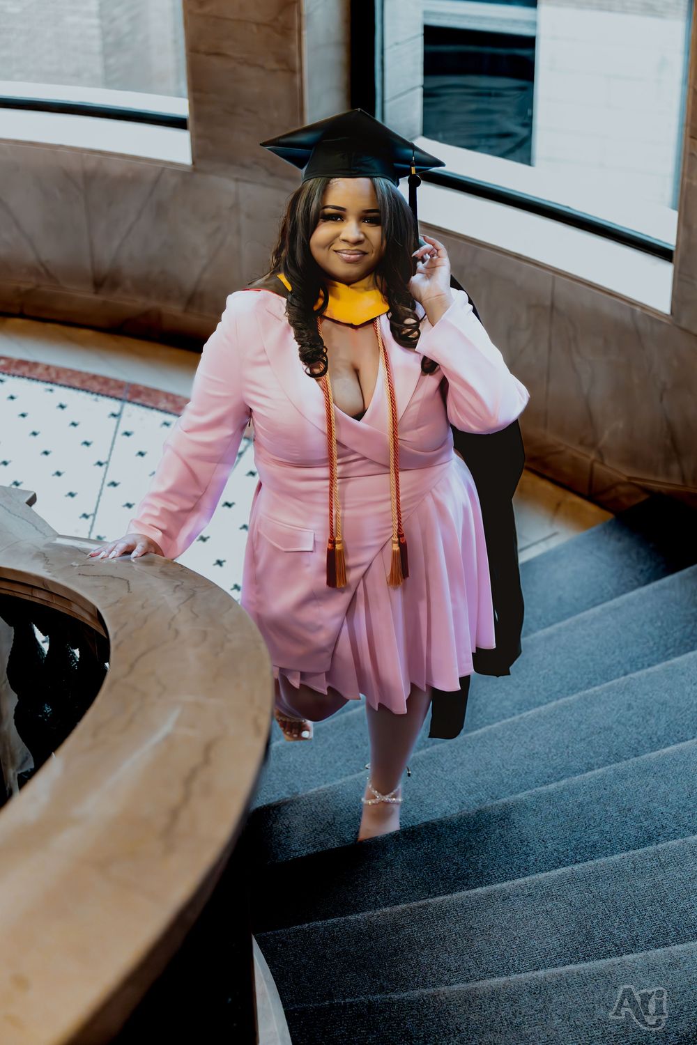 Young woman in pink dress and graduation cap and gown poses on elegant staircase during graduation photo session.