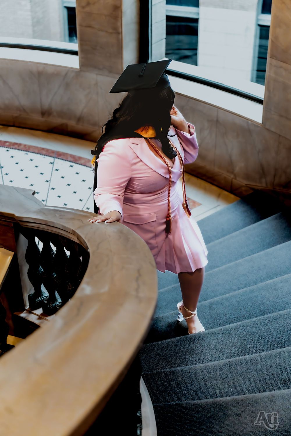 Graduate in pink suit and cap poses on curved staircase near large windows.