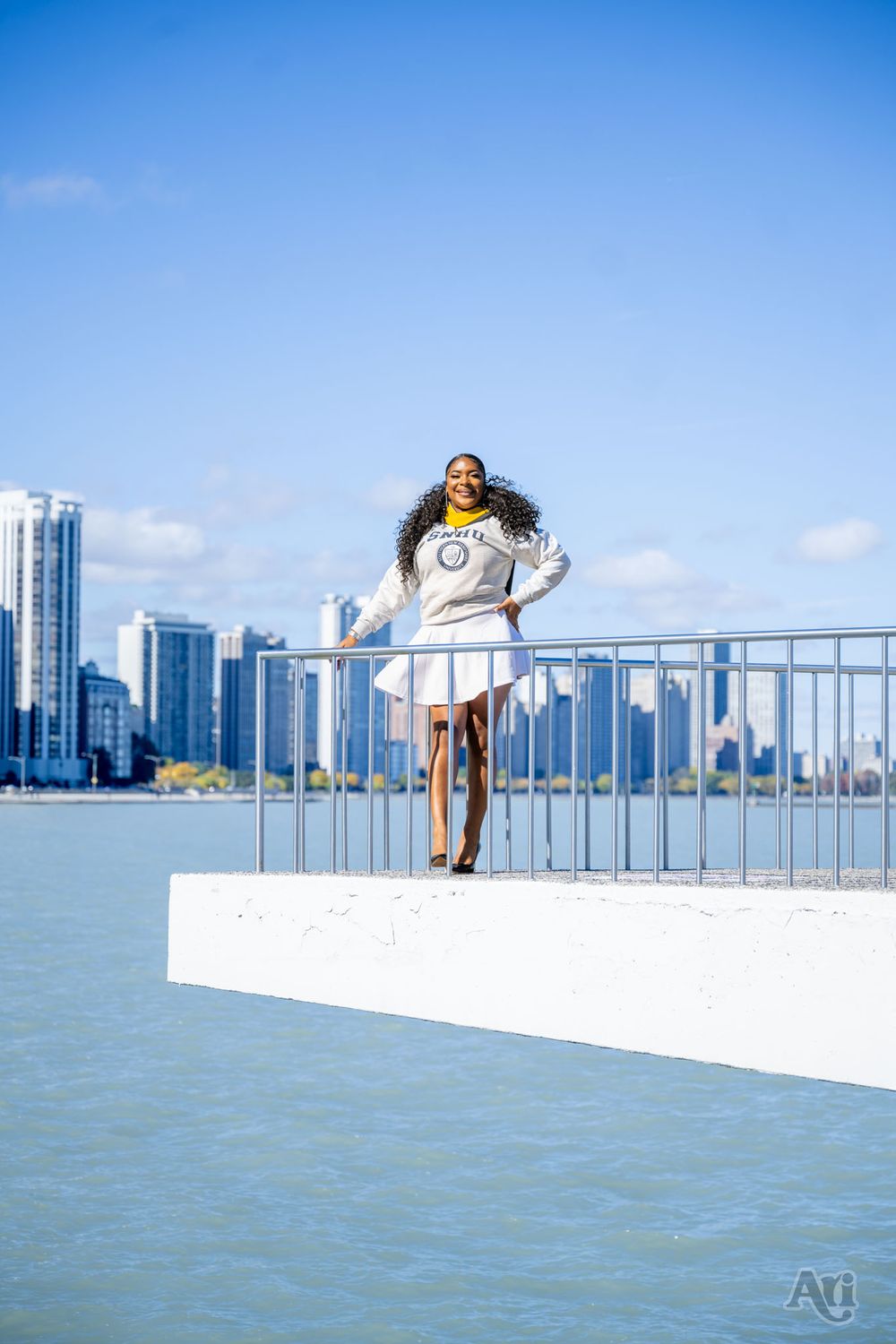 Person in white dress poses on waterfront balcony with Chicago skyline in background on sunny day.