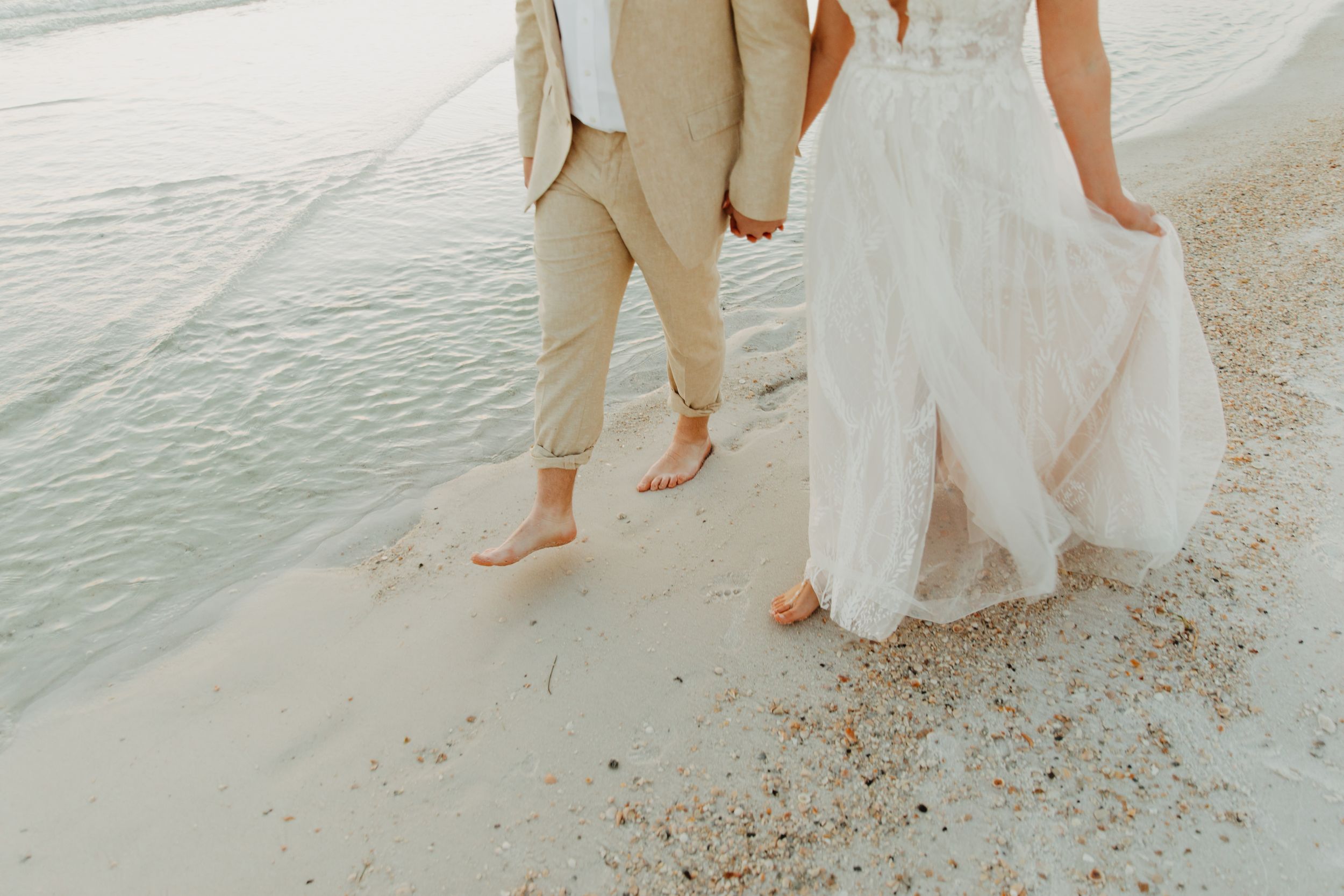 Barefoot couple walks along beach shoreline with groom in tan suit and bride in flowing white wedding dress.