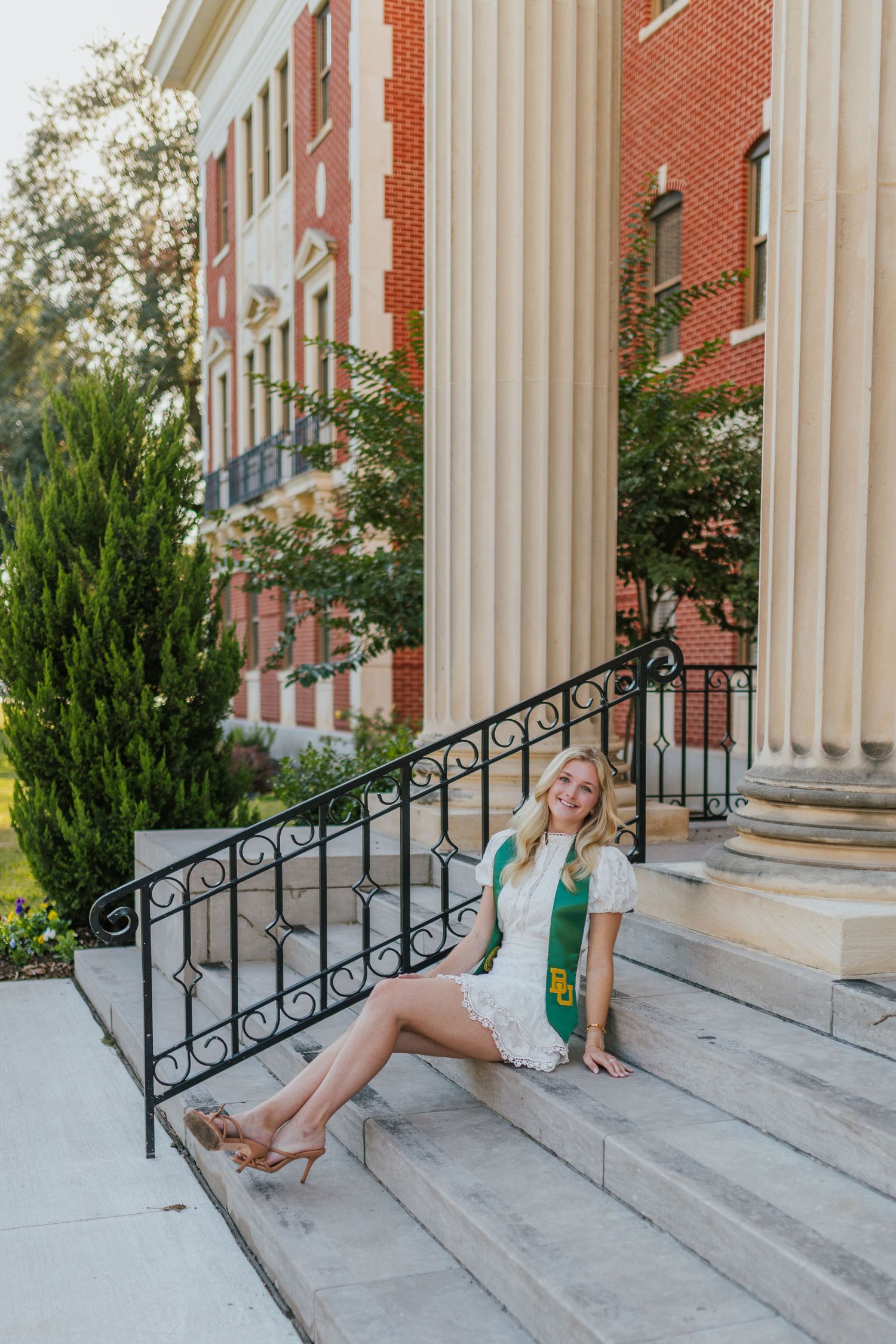 Blonde Baylor graduate in a white dress with a green BU stole sitting on steps beside tall columns.