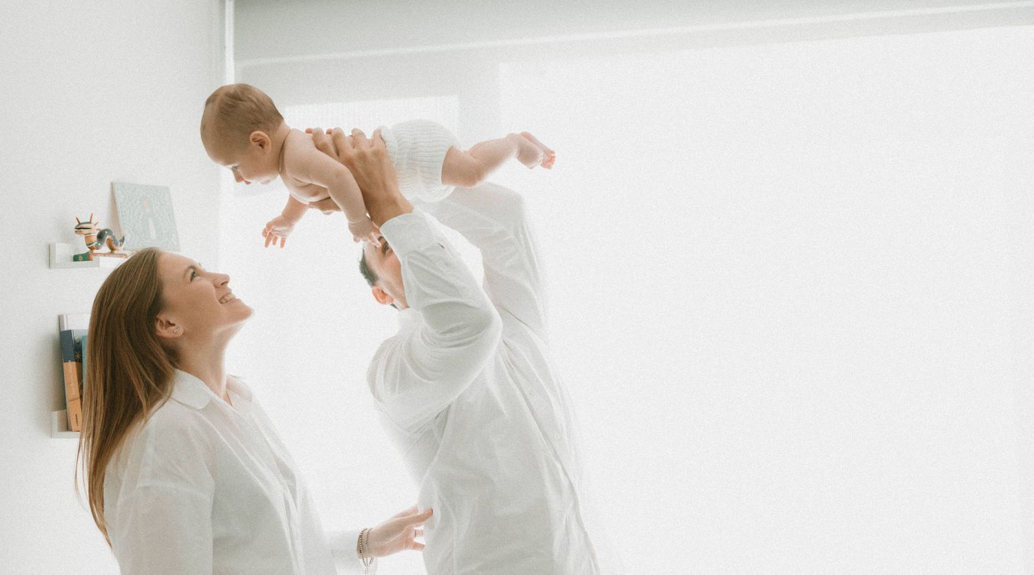 Parent lifting and playing with baby in white bright room.