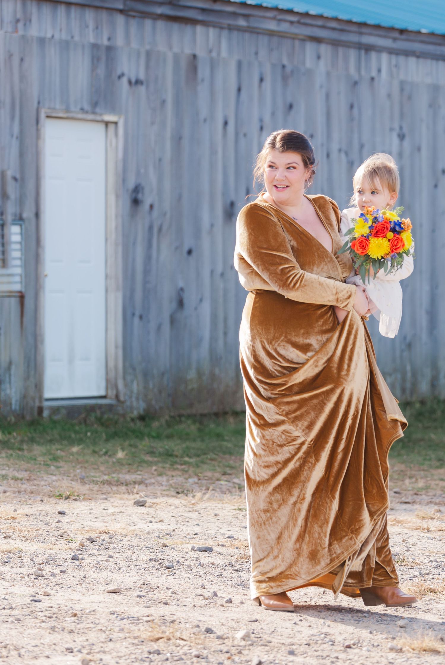 Person in velvet dress holding colorful autumn bouquet against rustic barn backdrop.