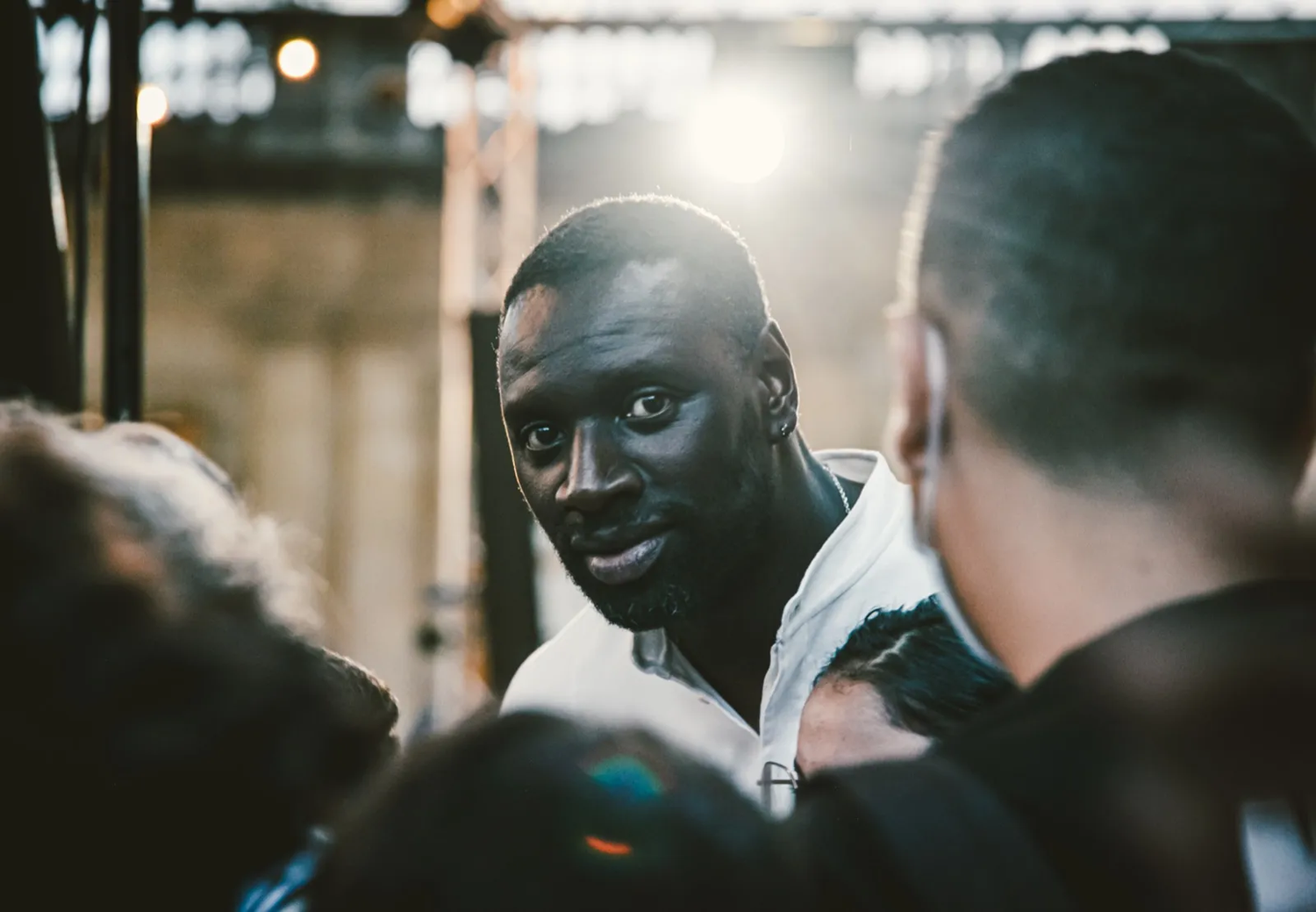 Homme au regard perçant tourné vers l’objectif, entouré de personnes floues dans une lumière rasante et dorée.