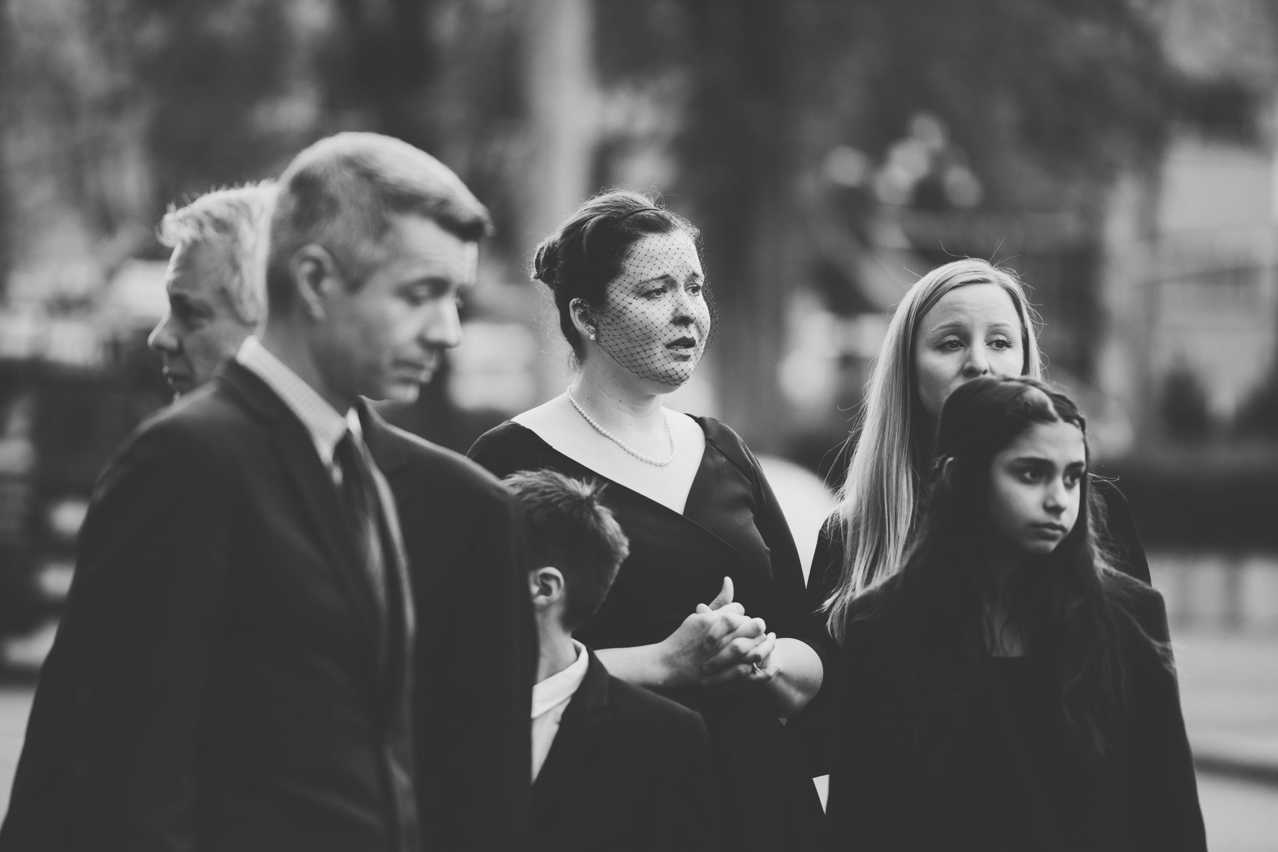 A black and white photograph shows a somber group of people dressed in dark formal attire walking together outdoors.