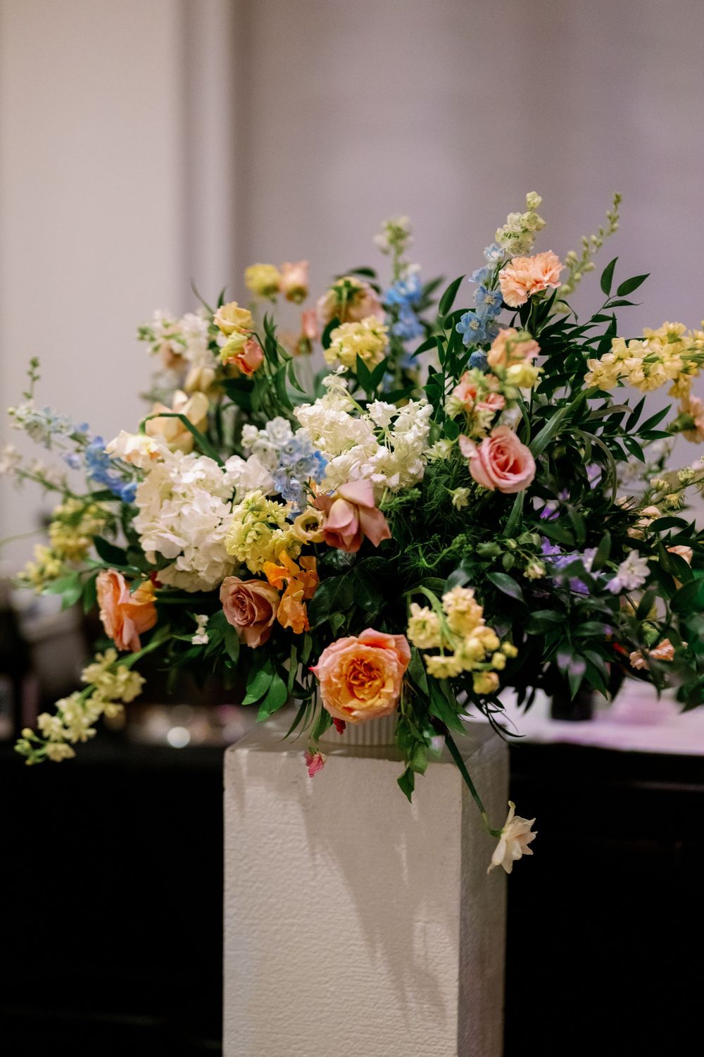 Elegant floral arrangement with peach roses, white blooms, and greenery on a white pedestal for a wedding.