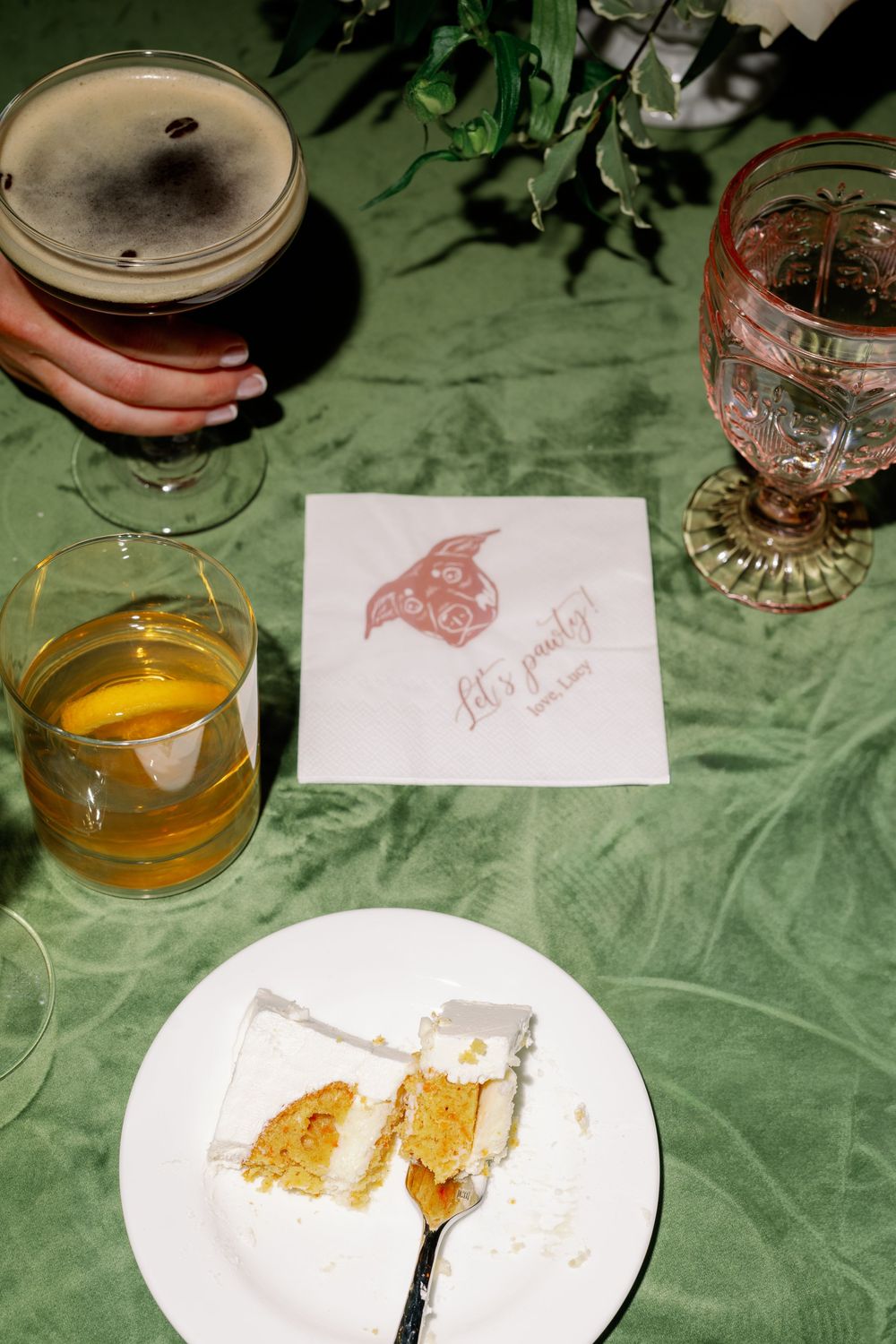 A slice of white cake on a plate beside drinks and a decorative napkin on a green tablecloth at a party.