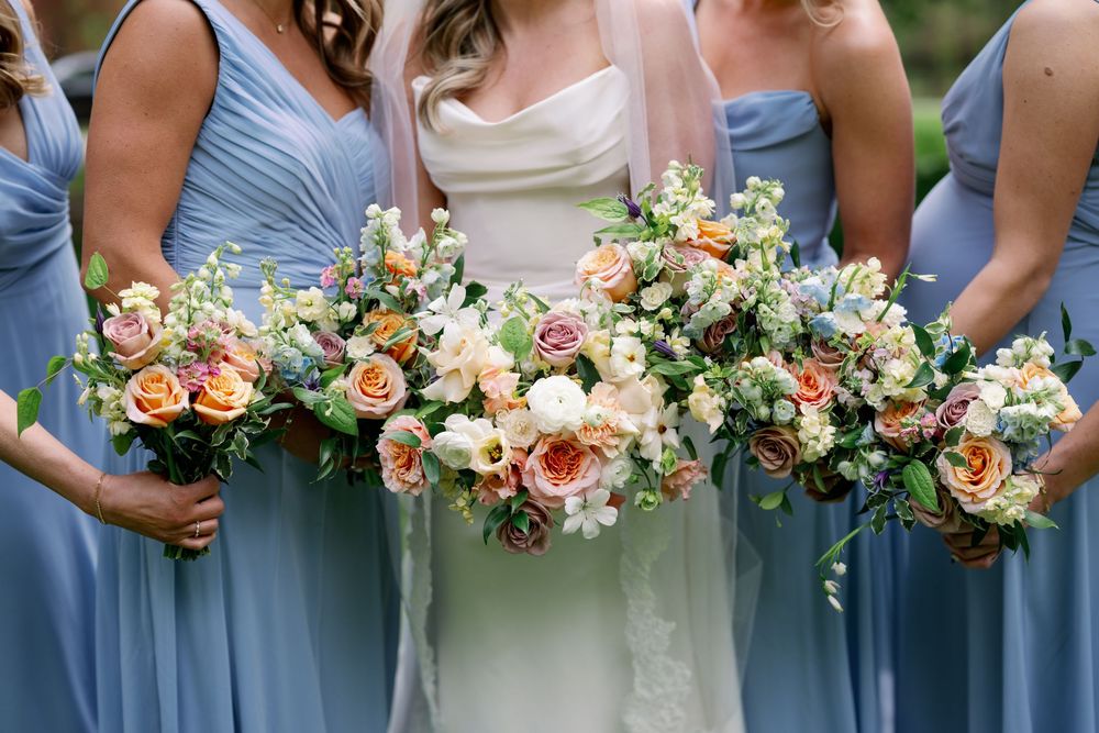 Bride in white gown and bridesmaids in blue dresses holding lush peach, blush, and ivory floral bouquets.