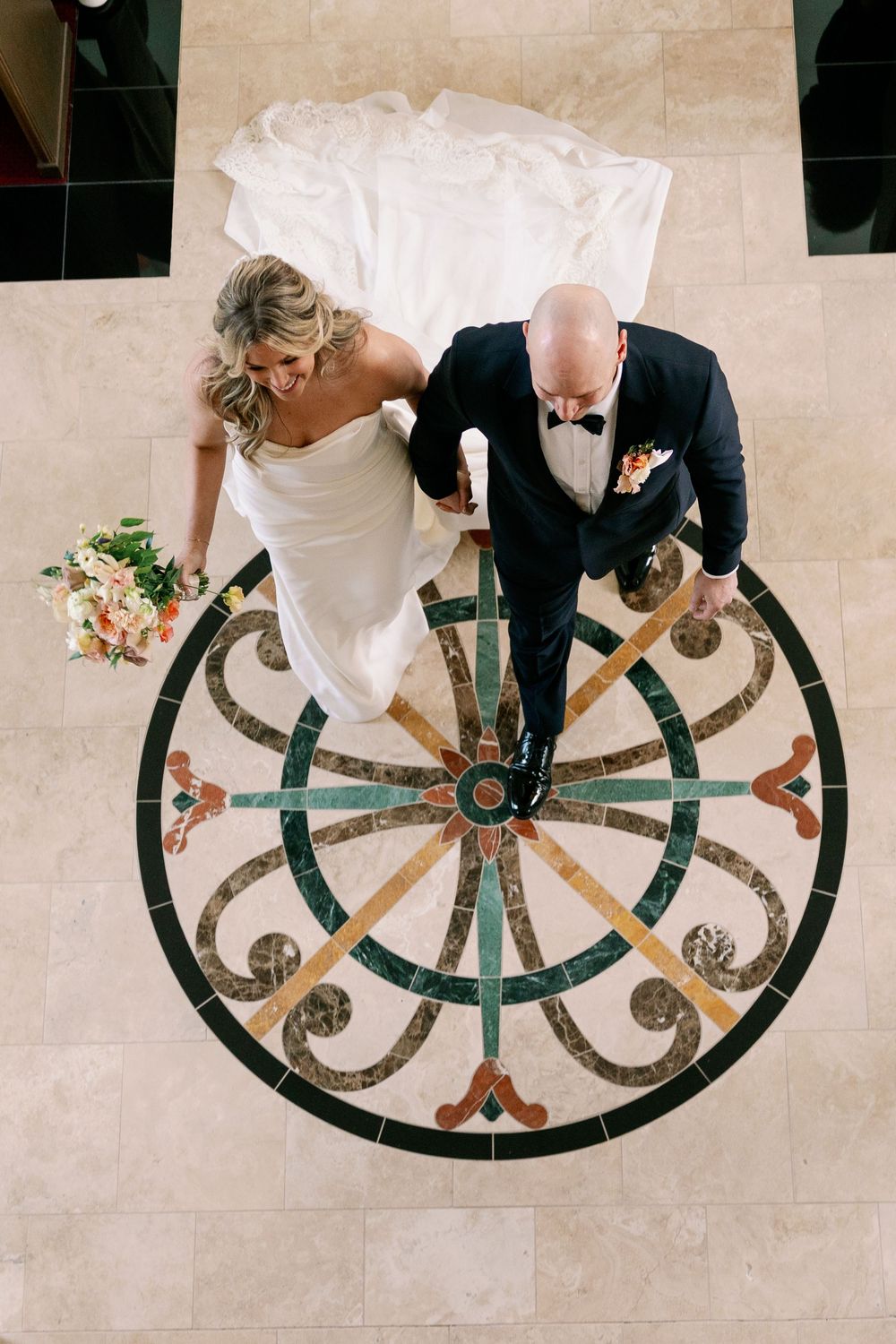 Aerial view of bride and groom walking on ornate mosaic floor medallion, bride holding bouquet.