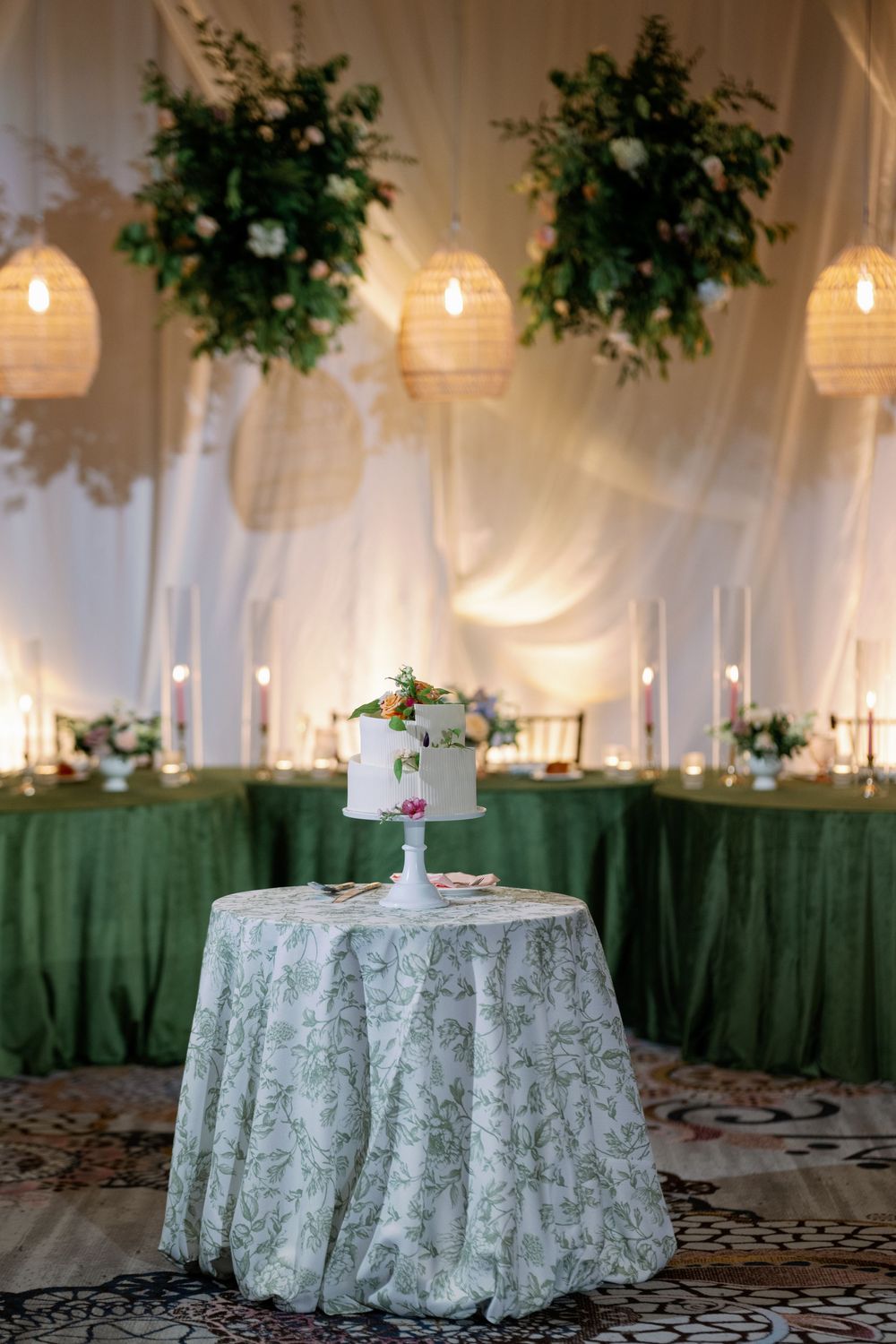 Elegant wedding reception with a two-tier white cake on a floral tablecloth, green draping, hanging florals, and candles.