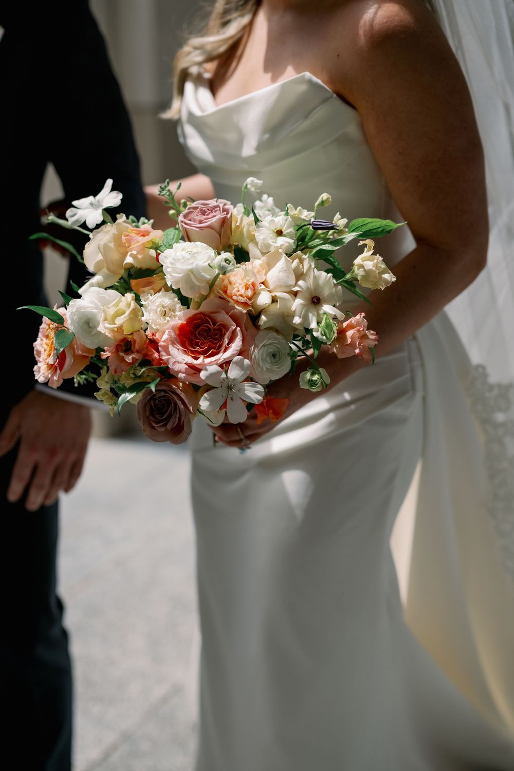 Bride in white strapless gown holds lush bouquet of peach, coral, and white roses with greenery on wedding day.