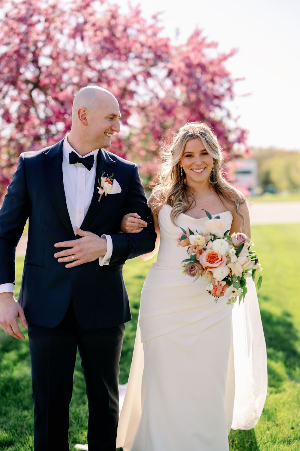 Smiling bride and groom walk arm in arm before pink cherry blossom trees on their wedding day.