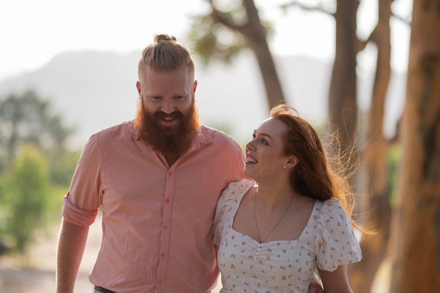A romantic couple embraces while overlooking a scenic mountain landscape with rolling hills and valleys at sunset.