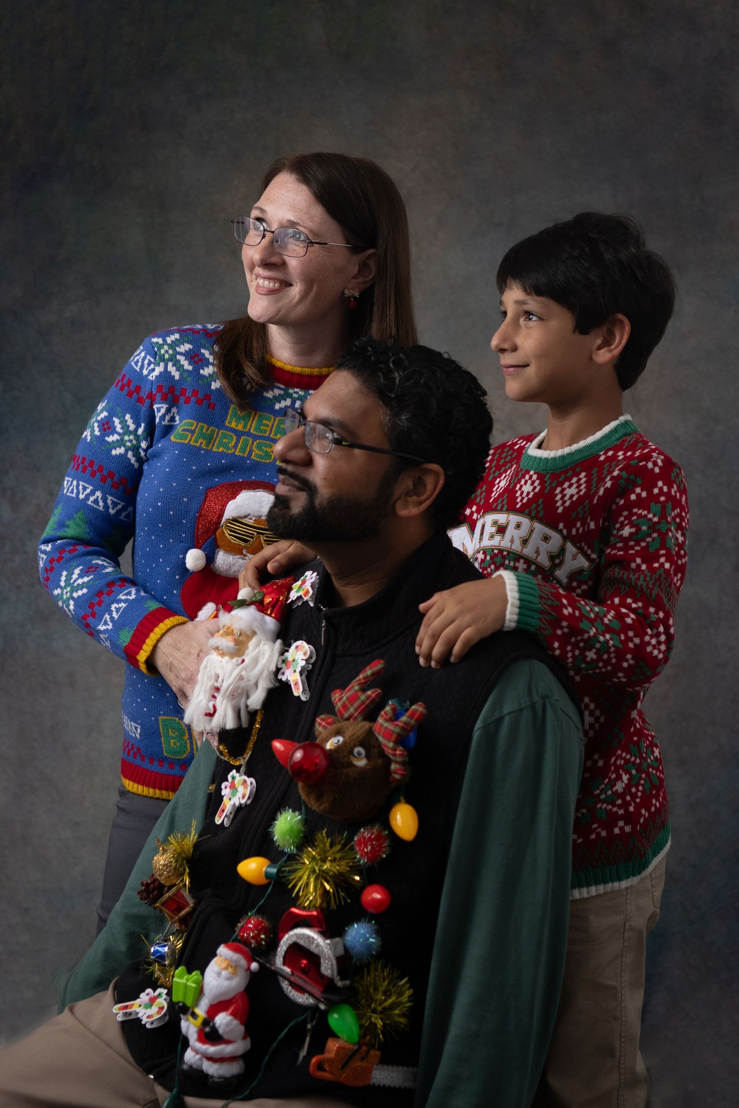 Two people wearing festive holiday sweaters pose together with one adorned in Christmas decorations and Santa figures.