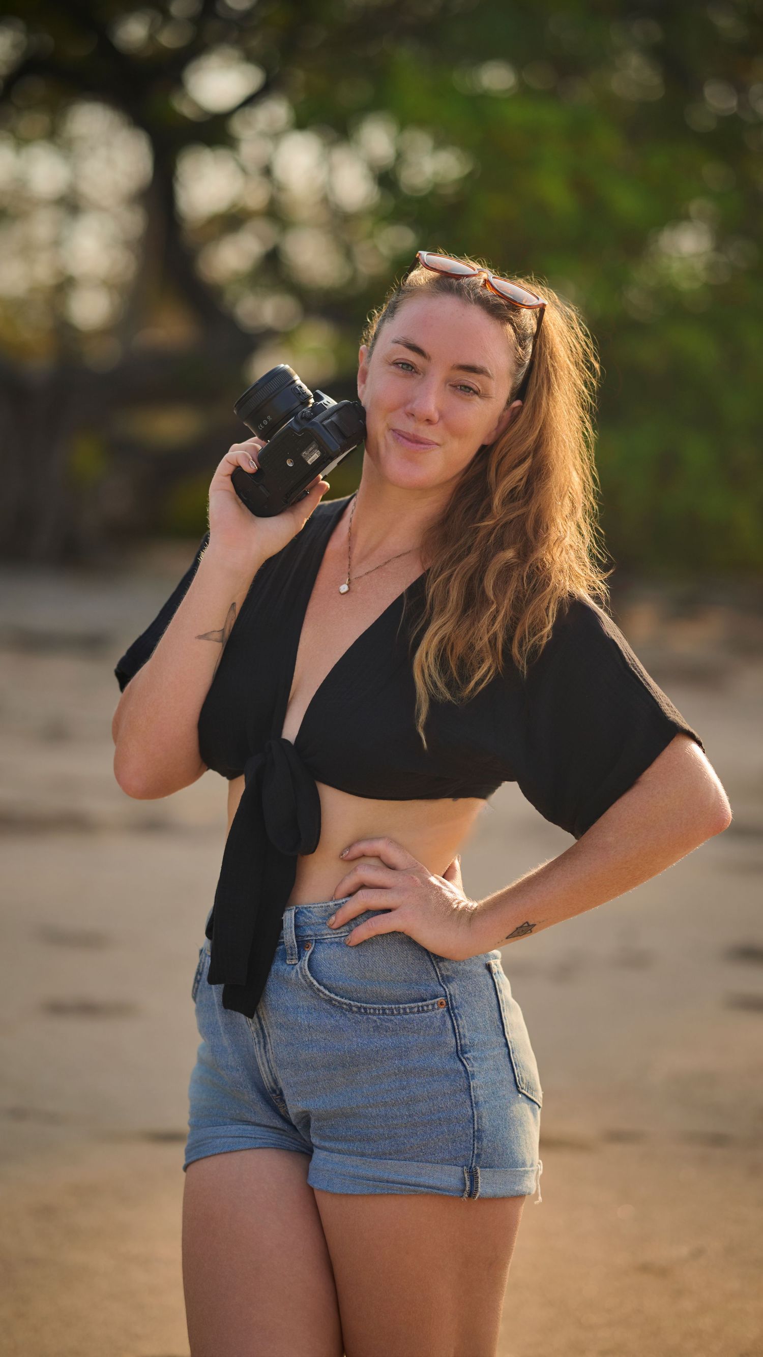 A person in denim shorts and black top poses with a camera in a series of outdoor beach sunset photos.