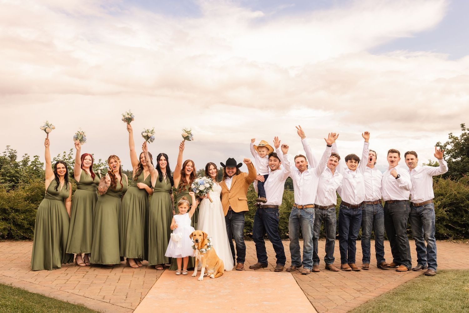 Wedding party celebrates with raised arms outdoors, bridesmaids in sage green dresses and groomsmen in white shirts and jeans.