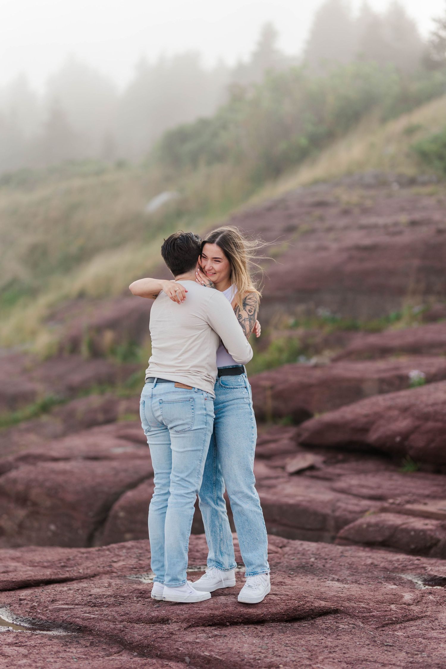 Happy couple embracing on rugged red coastal rocks of Flatrock, Newfoundland with misty scenery.