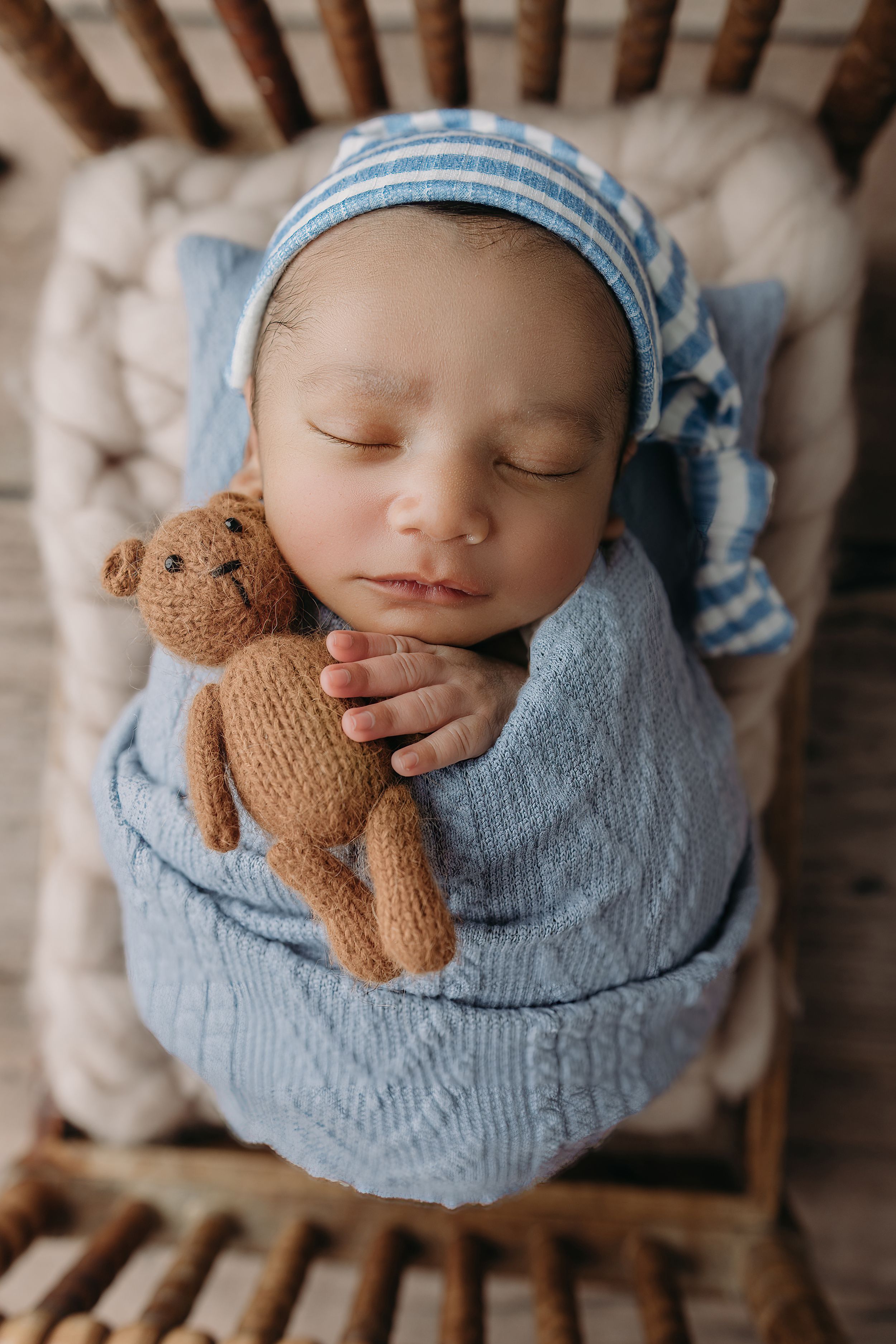 Sleeping newborn wrapped in soft blue knit blanket holding small brown teddy bear in professional portrait series.
