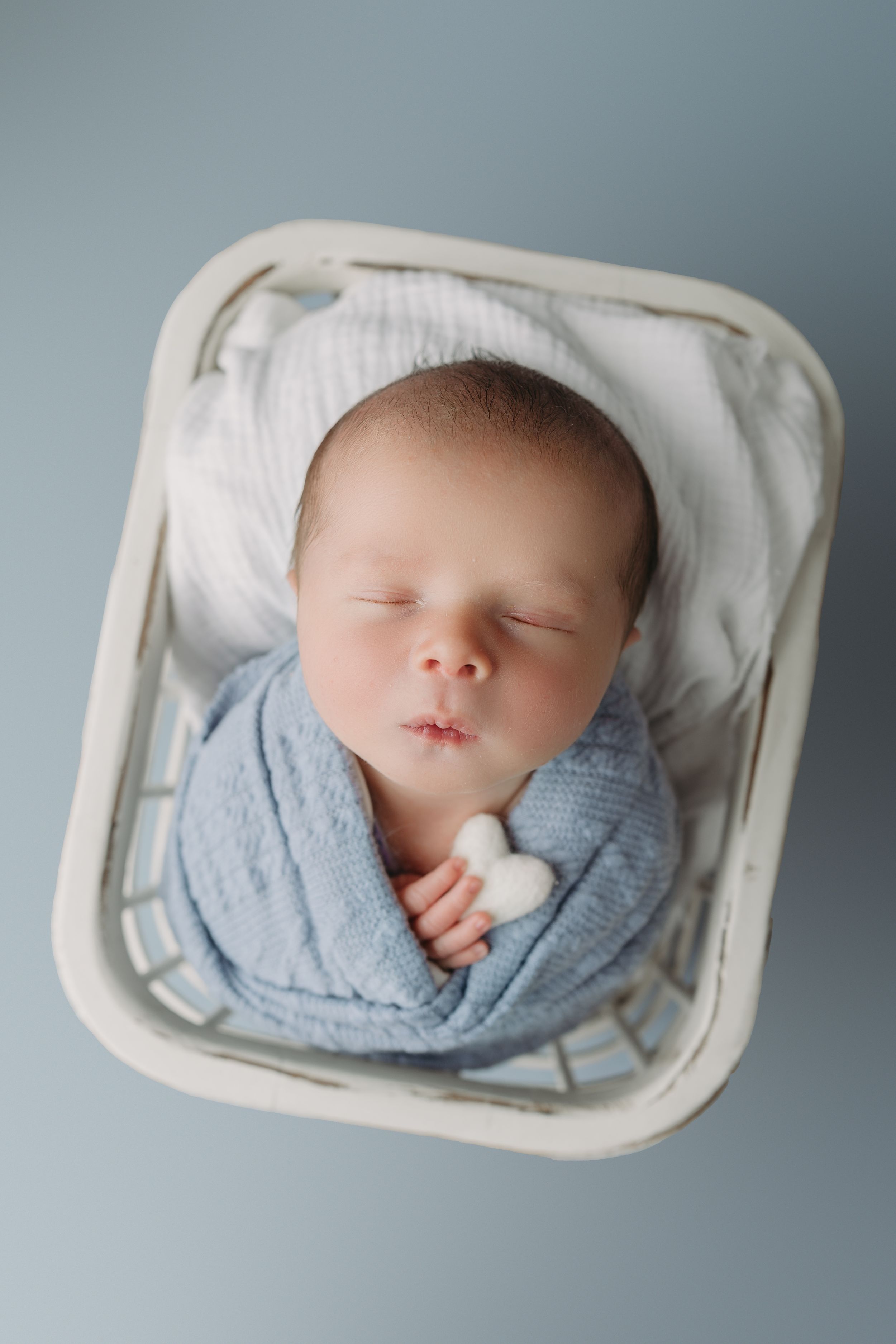 Sleeping newborn wrapped in soft blue blanket lying in white basket on grey background.