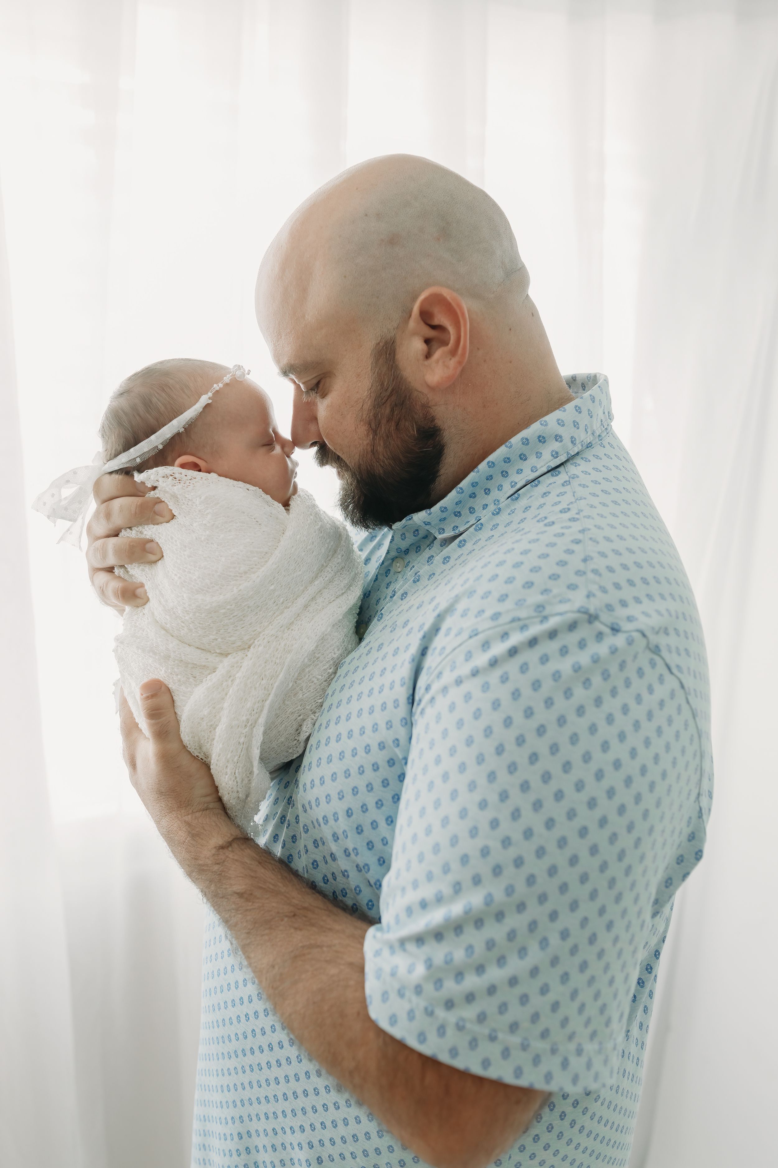A tender moment between an adult in a blue polka dot shirt and a swaddled newborn against a light curtained background.