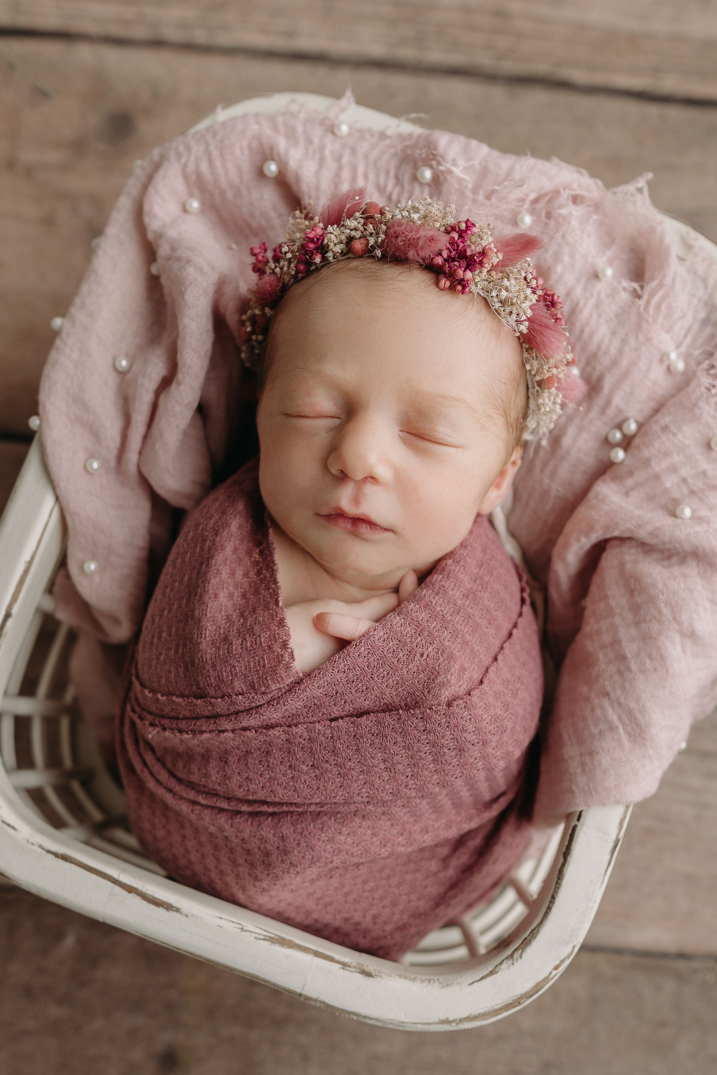 Sleeping newborn swaddled in mauve wrap with delicate floral crown lying on soft pink fabric in white basket.