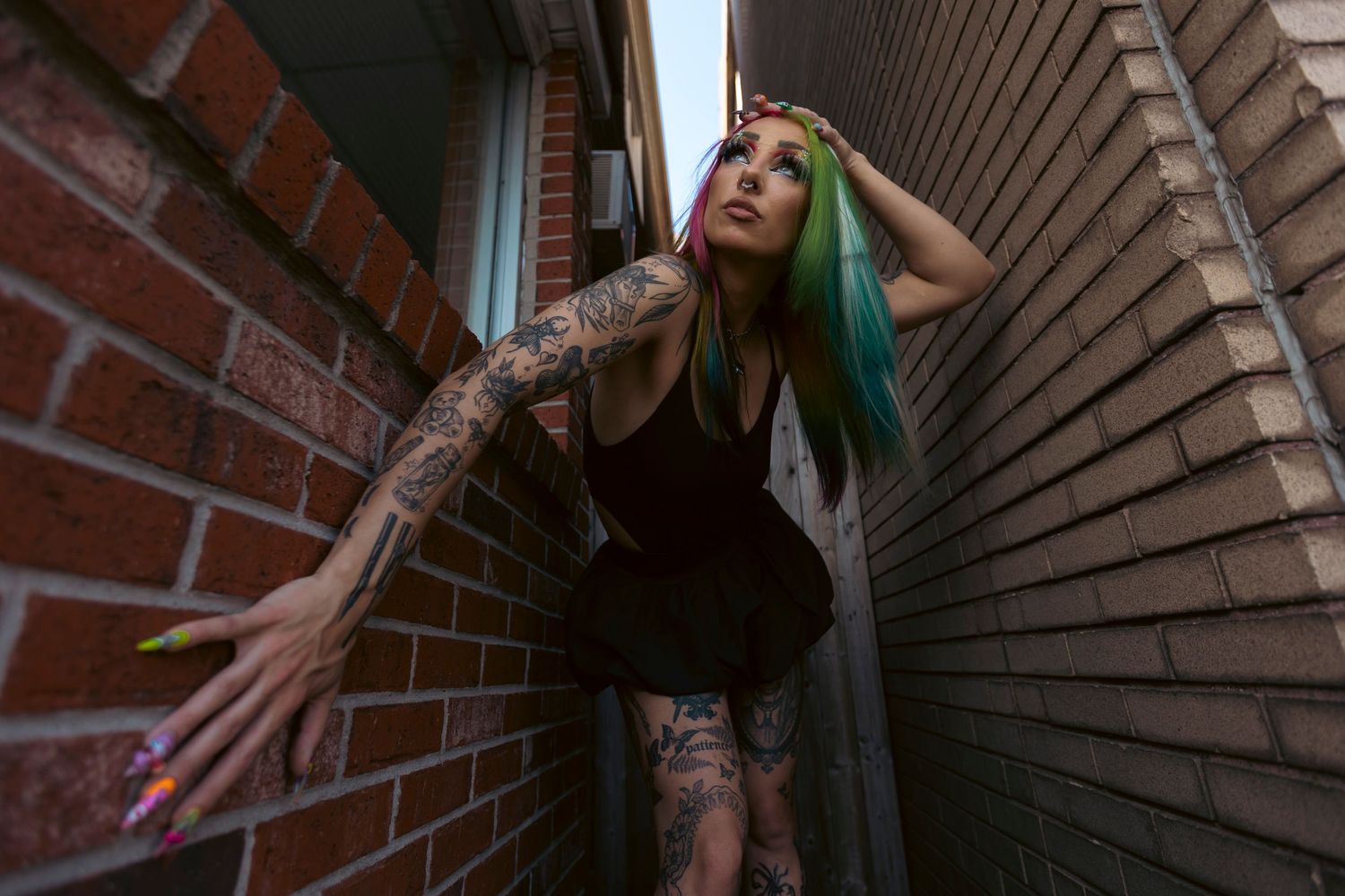 Person with bright green hair and tattoos poses between brick walls in dark moody lighting.