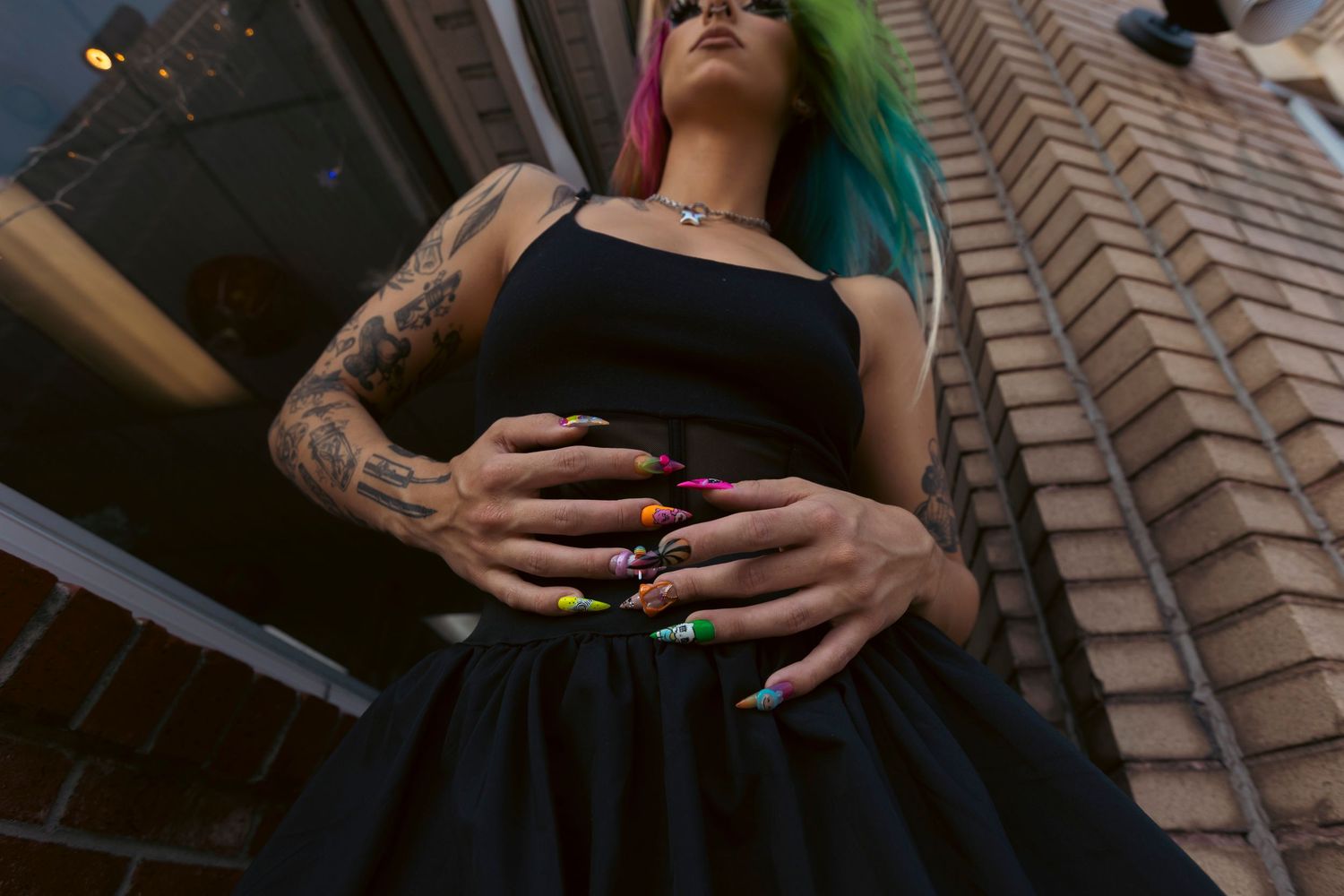 Woman in black dress with colorful hair and arm tattoos poses against brick wall, showing painted fingernails.